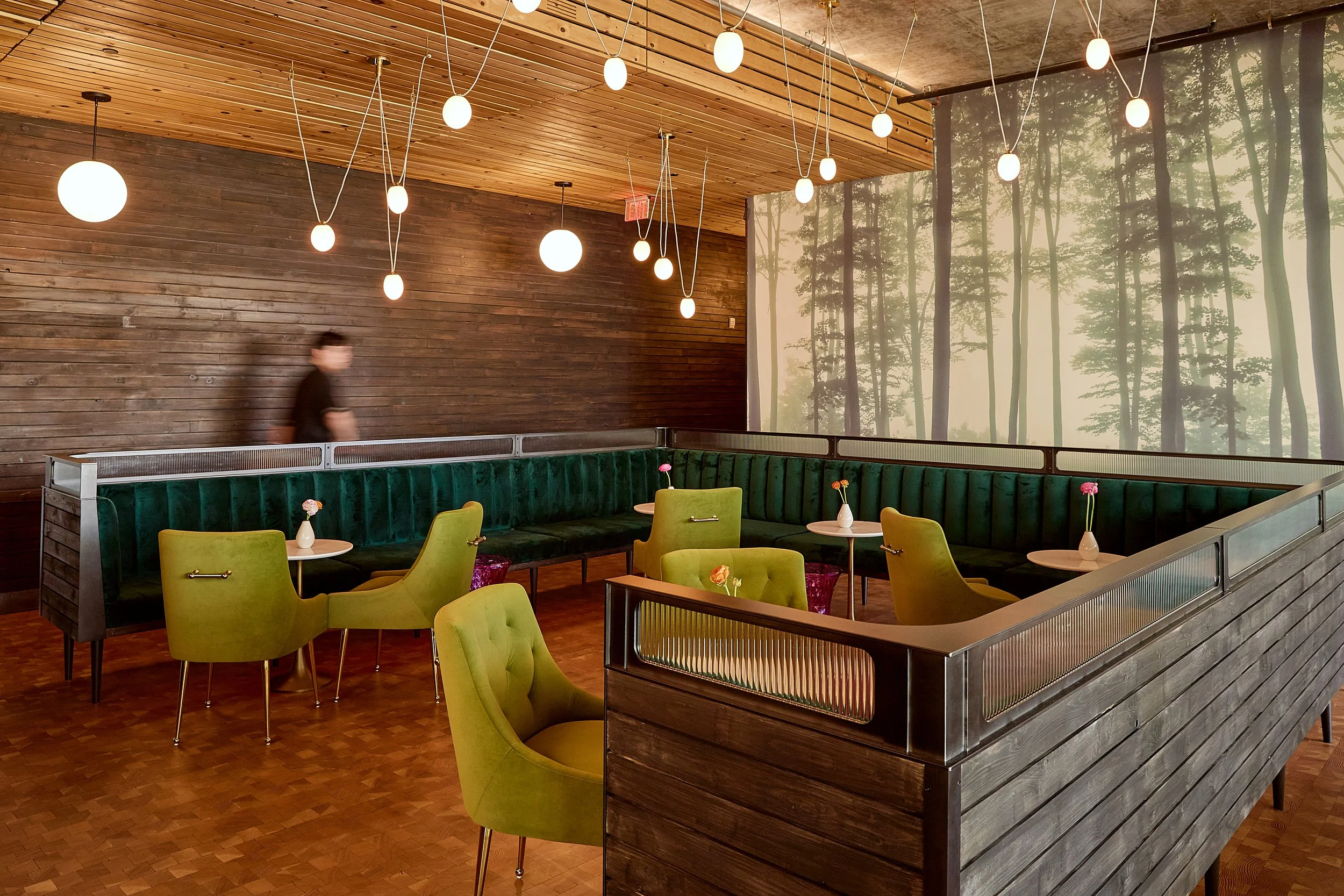 Interior of a restaurant or cafe with a wooden wall and ceiling, green upholstered seating, lime green chairs, small tables with vases of pink flowers, and hanging round white lights. A person is blurred, walking past in the background near the wall,