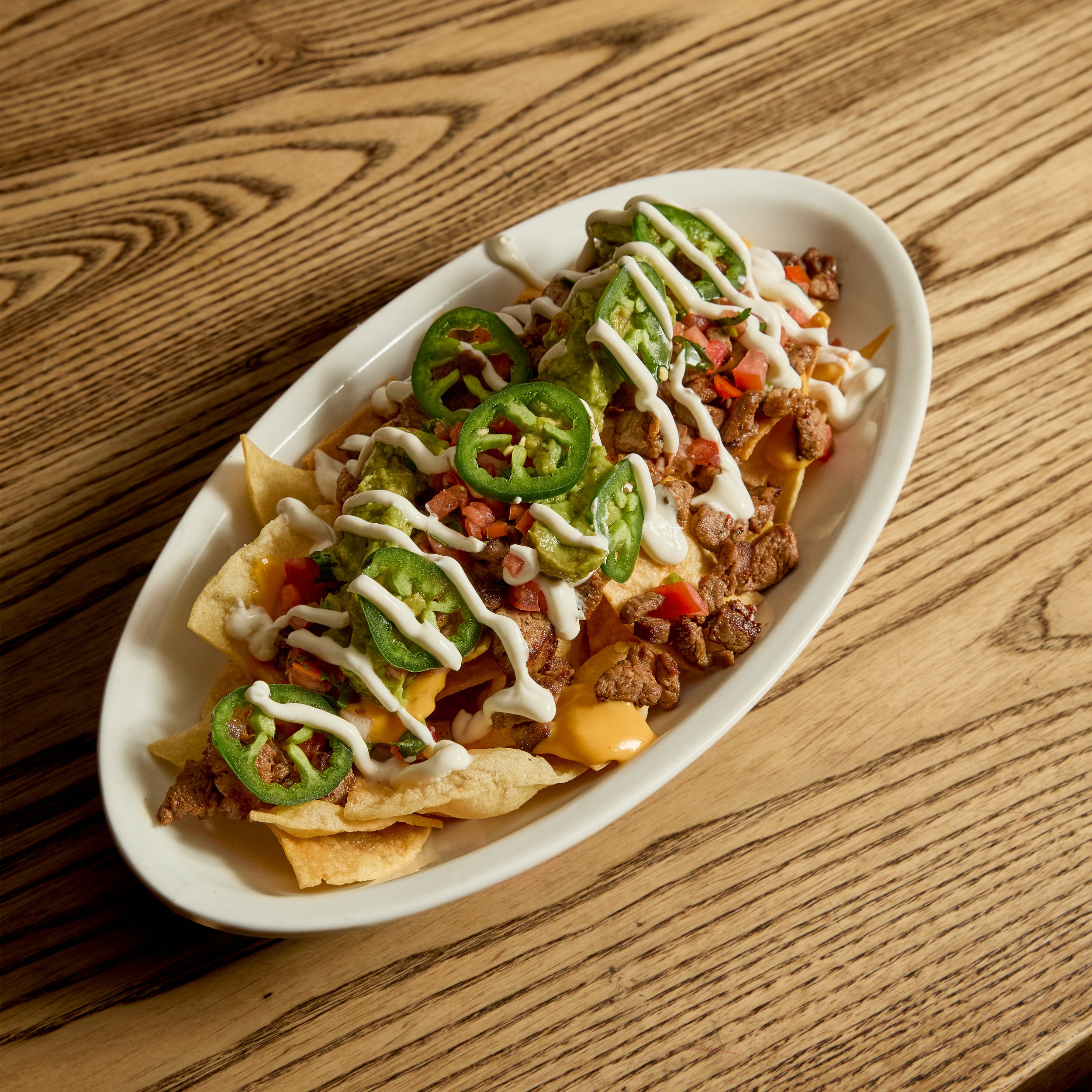 A plate of crunchy beef tacos topped with sliced jalapeños, shredded cheese, sour cream, and diced tomatoes on a wooden table.