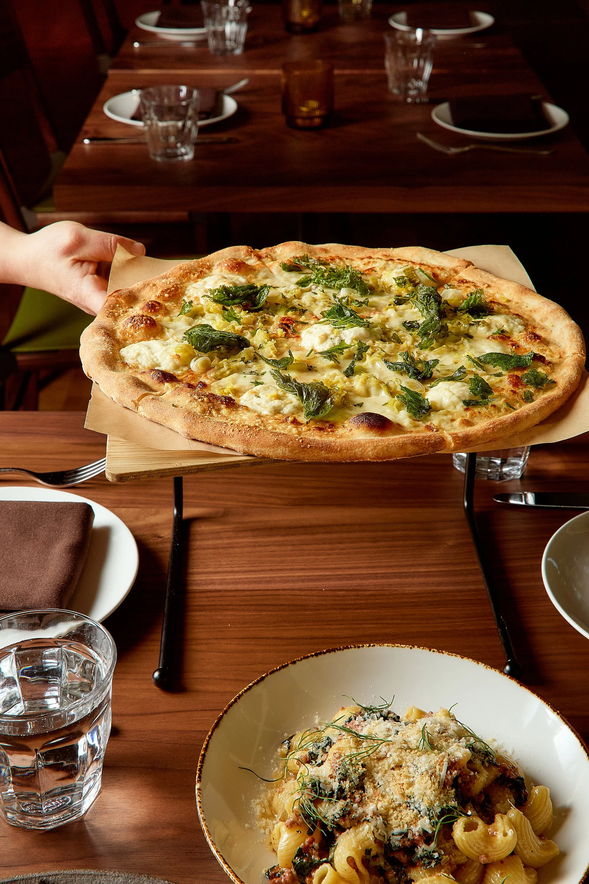 A hand holding a wooden tray with a pizza topped with green herbs and cheese, placed over a dining table in a restaurant setting.