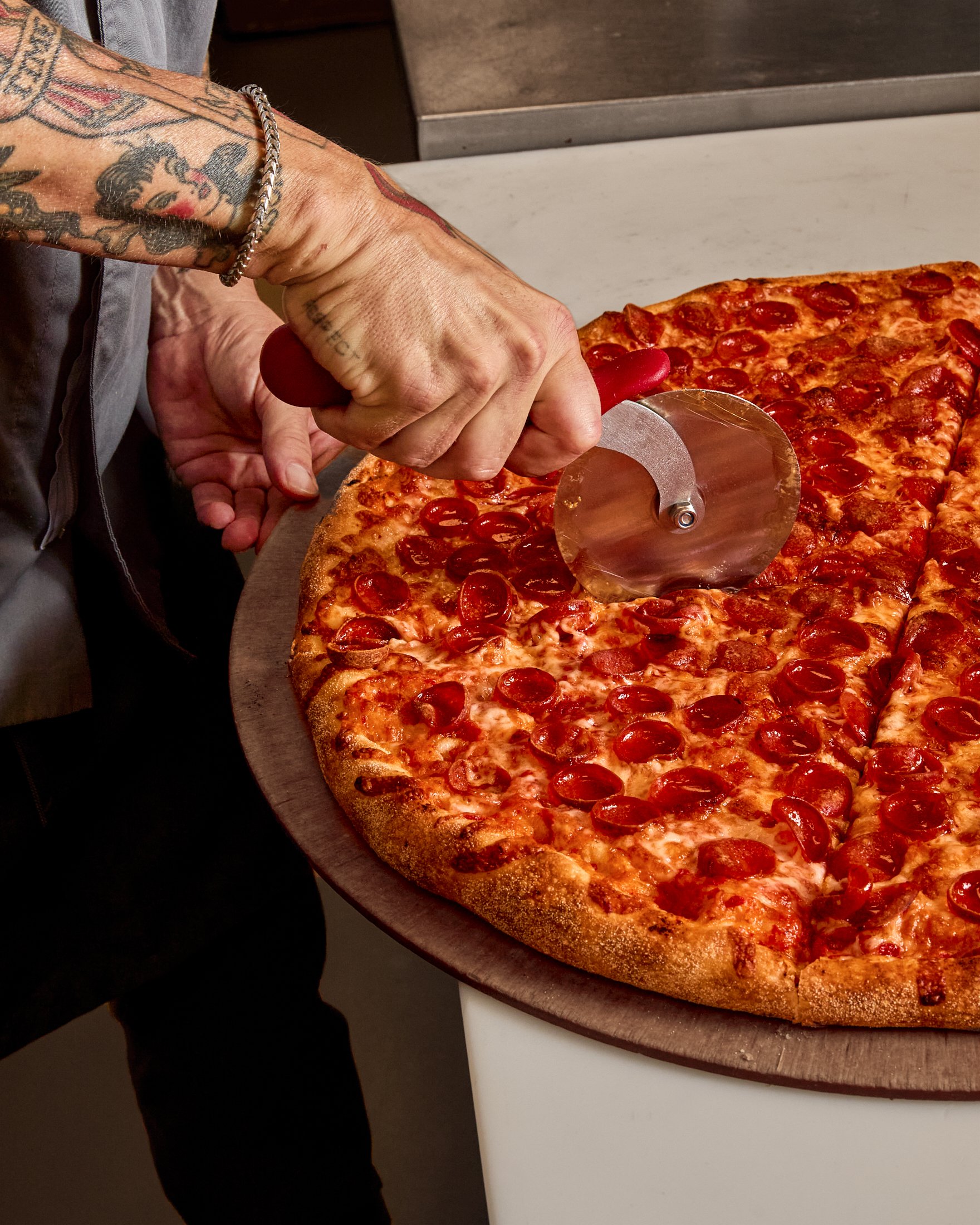 Close-up of a person slicing a large pepperoni pizza with a pizza cutter, showing the pizza's toppings and crust.