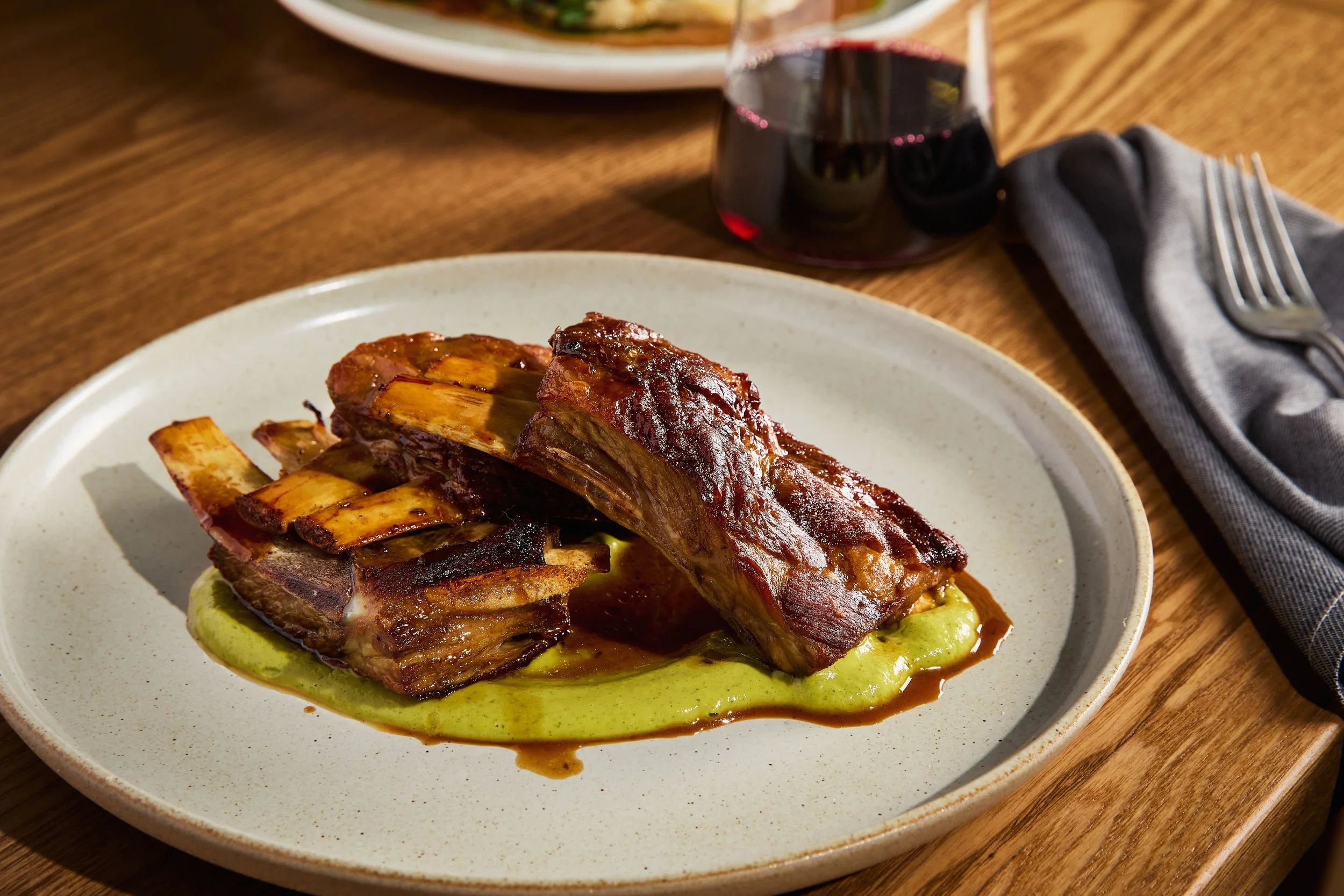Hearty beef short ribs served on a plate with mashed potatoes, topped with crispy bacon, with a glass of red wine on a wooden table.