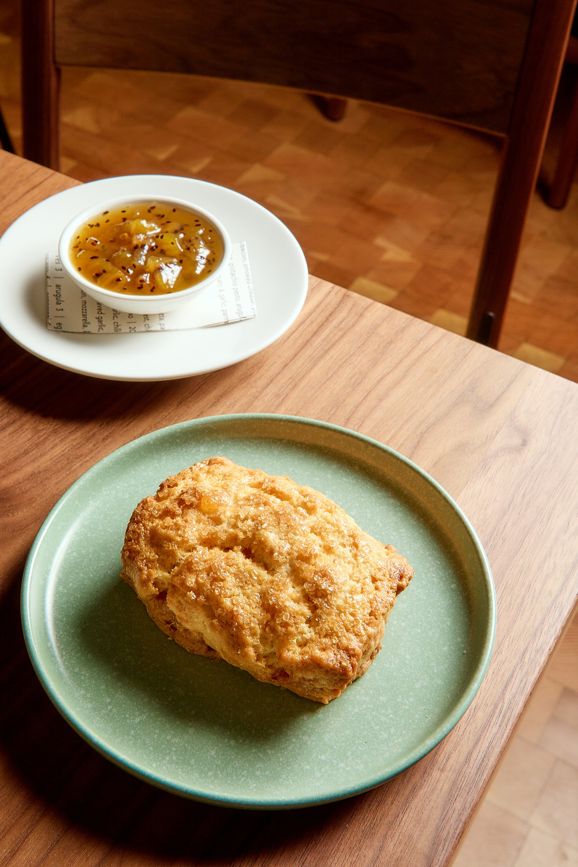 A green plate with a large, golden-brown fried food item on a wooden table. In the background, there is a small cup of sauce or jam on a white plate, sitting on a piece of paper with printed text.