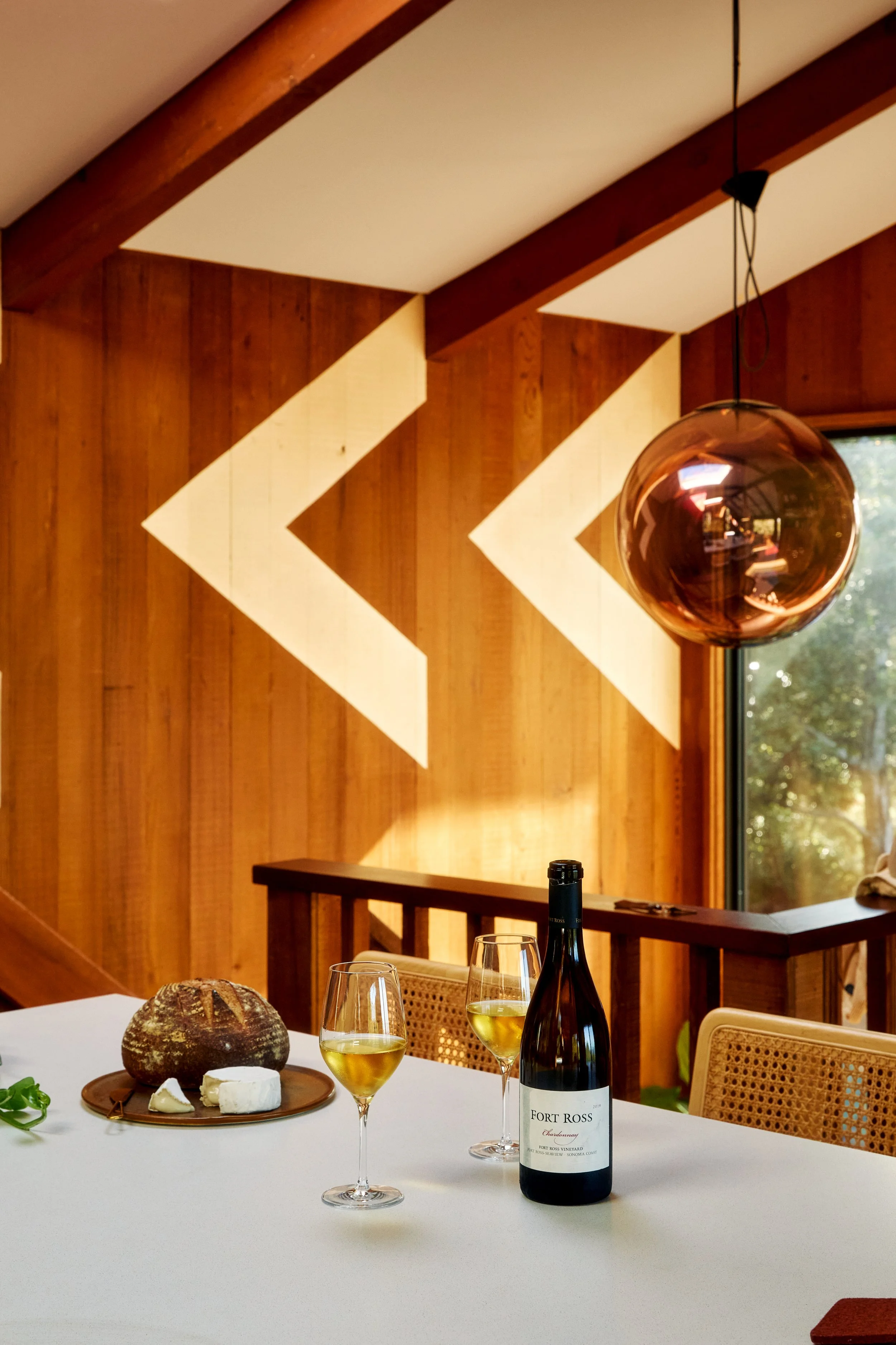 Interior of a dining room with a white tablecloth, a bottle of white wine, two glasses of white wine, a round cheese, and a loaf of bread on a wooden tray, with a wooden wall featuring white geometric shapes and a large glass pendant light.