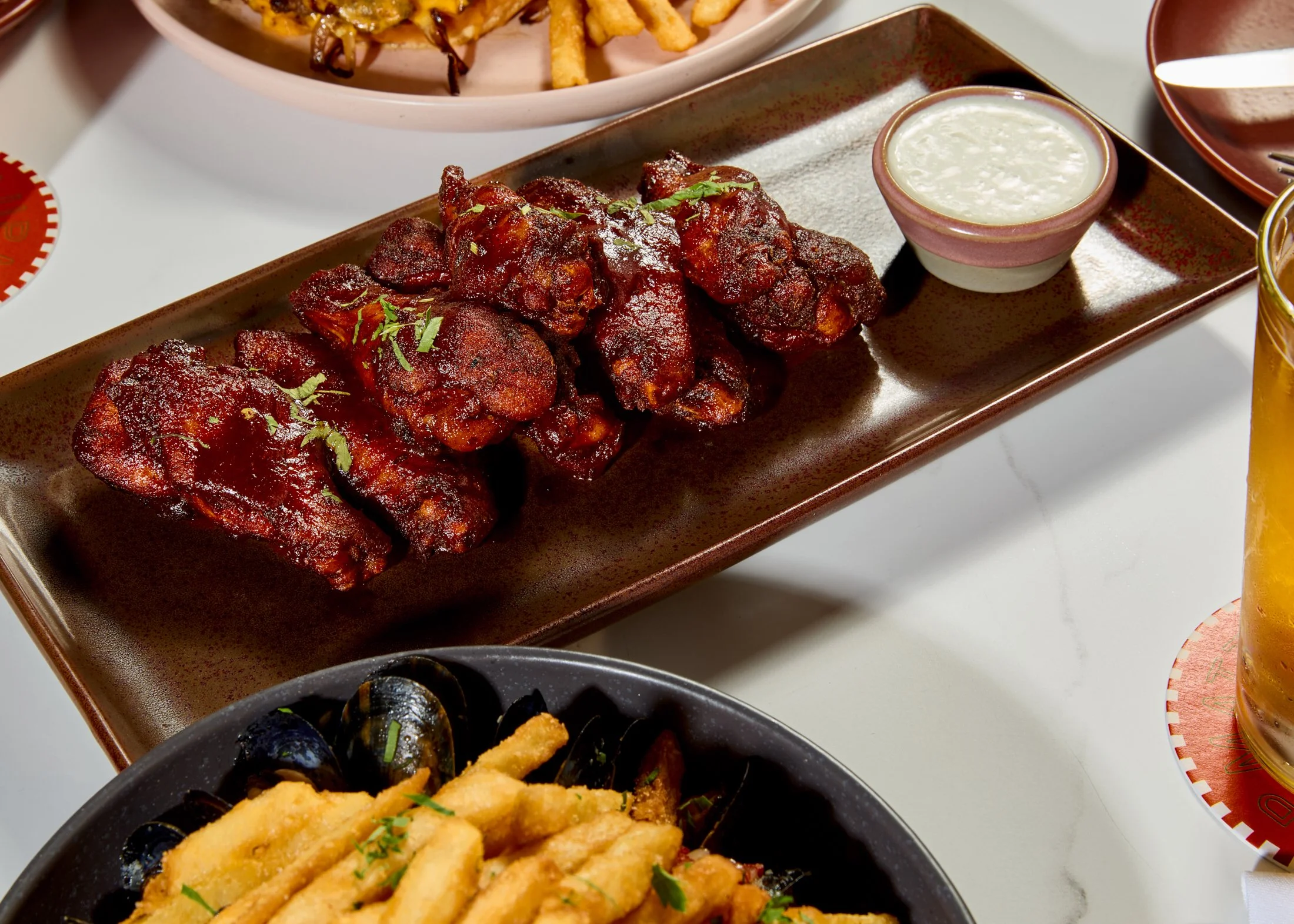 Plate of crispy glazed chicken wings garnished with chopped green herbs, served with a side of white dipping sauce. Surrounding dishes include french fries, jalapeño poppers, and a bowl of rice pudding, set on a table with a beverage.