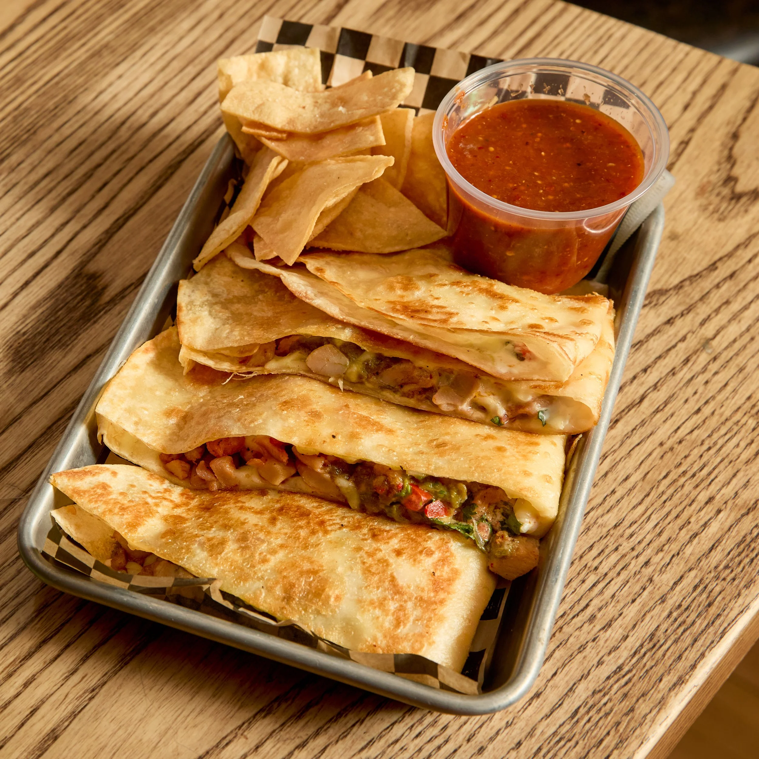 A metal tray with four quesadillas, a cup of red sauce, and some potato chips on a wooden table.