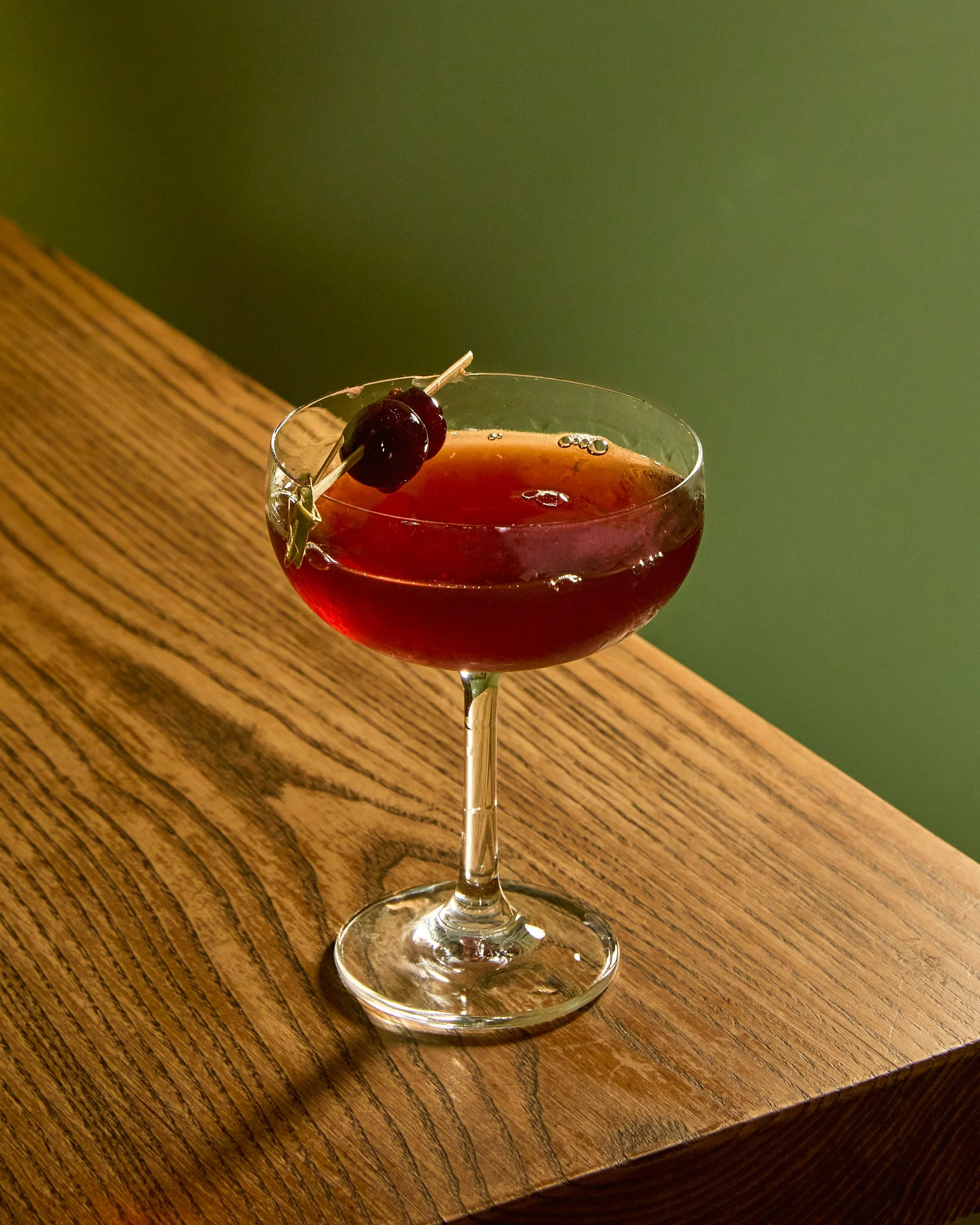 A cocktail glass with a red drink garnished with a cherry on a skewer, sitting on a wooden surface.