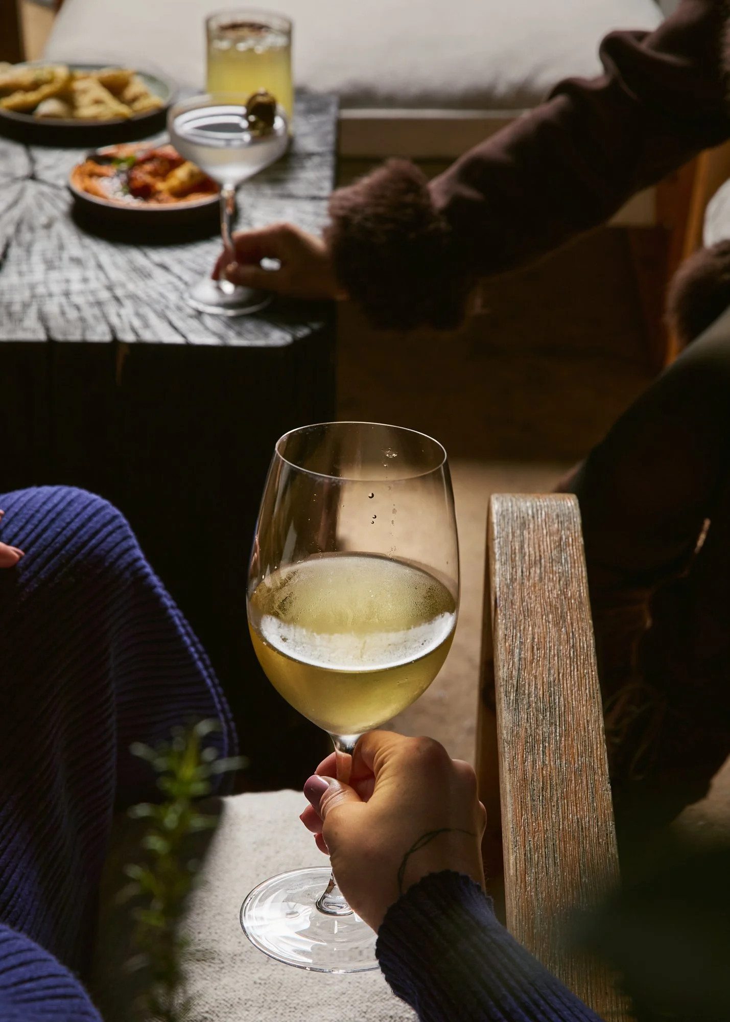 Person holding a glass of white wine in front of a table with food and drinks