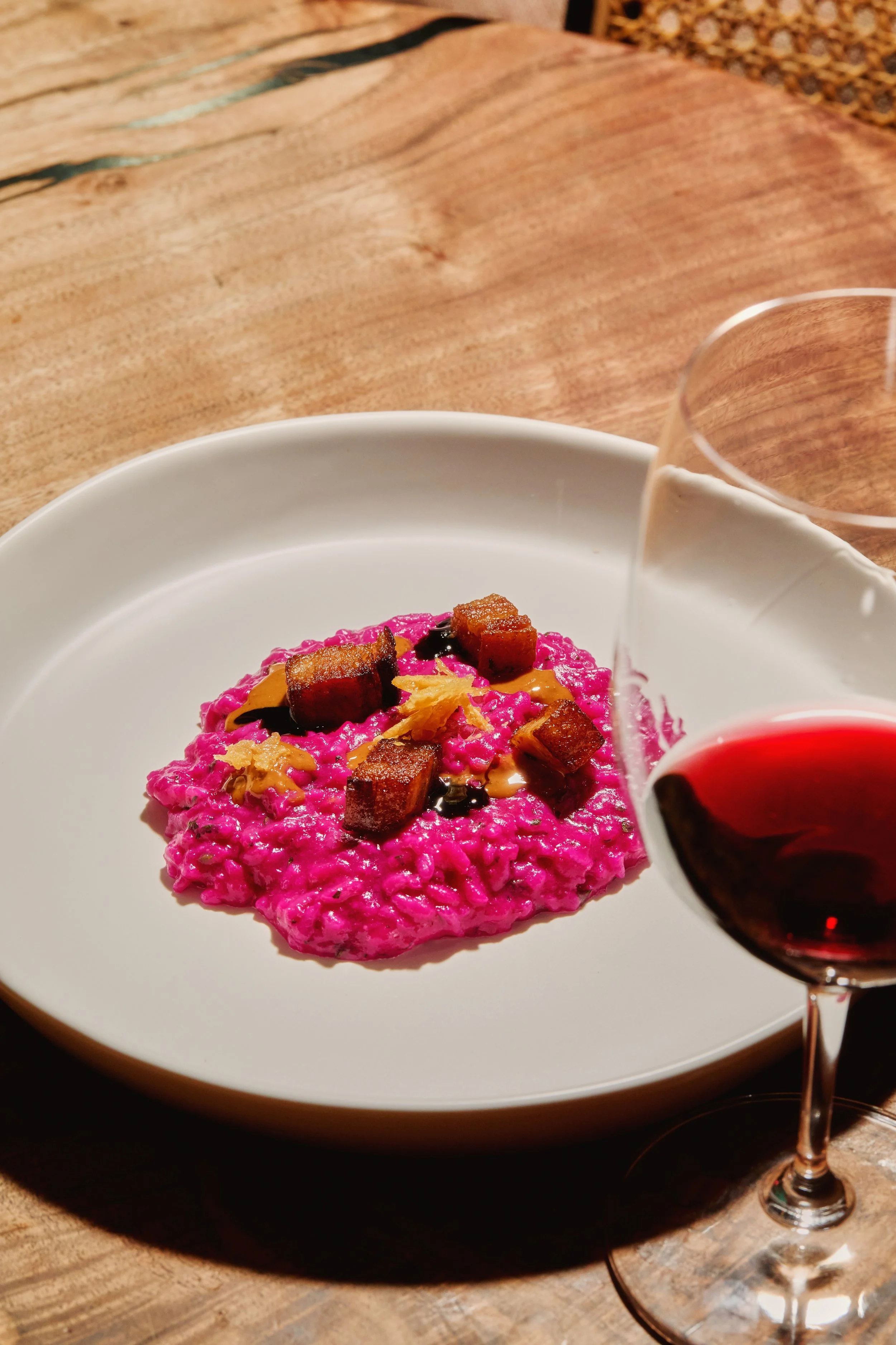 A white plate with a serving of vibrant purple rice topped with crispy tofu cubes and garnishes, with a glass of red wine nearby, on a wooden table.