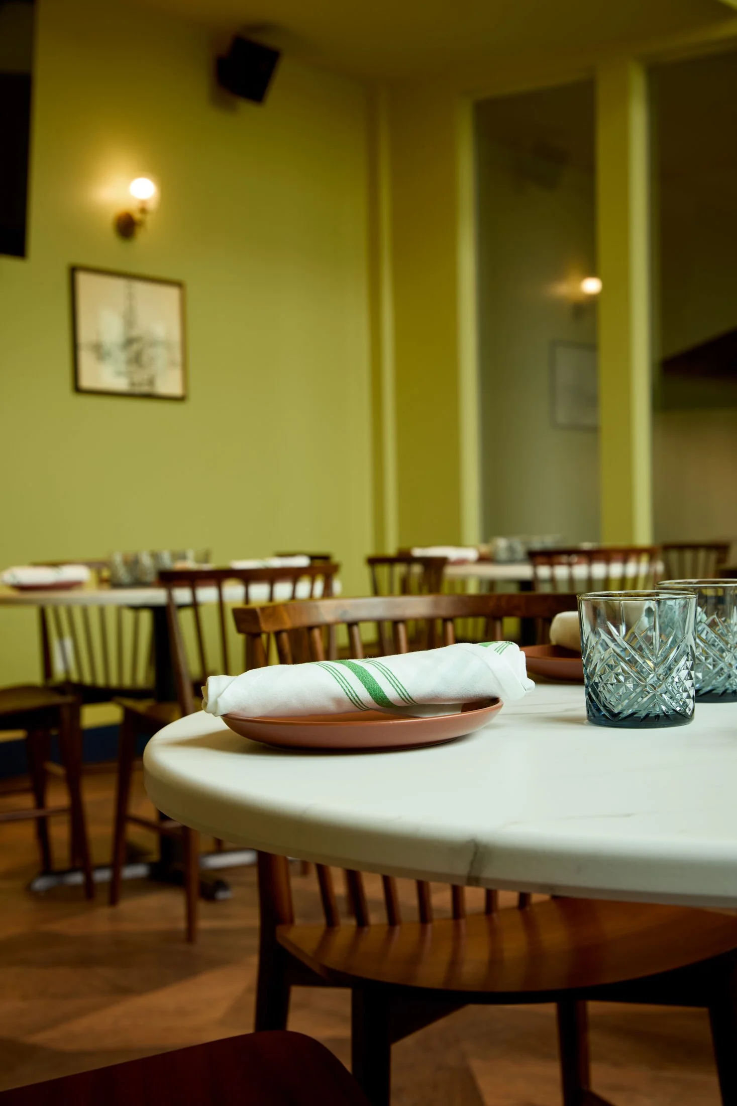 A restaurant dining area with round tables, chairs, napkins, glassware, and a yellow and green wall with framed pictures and wall-mounted lamps.