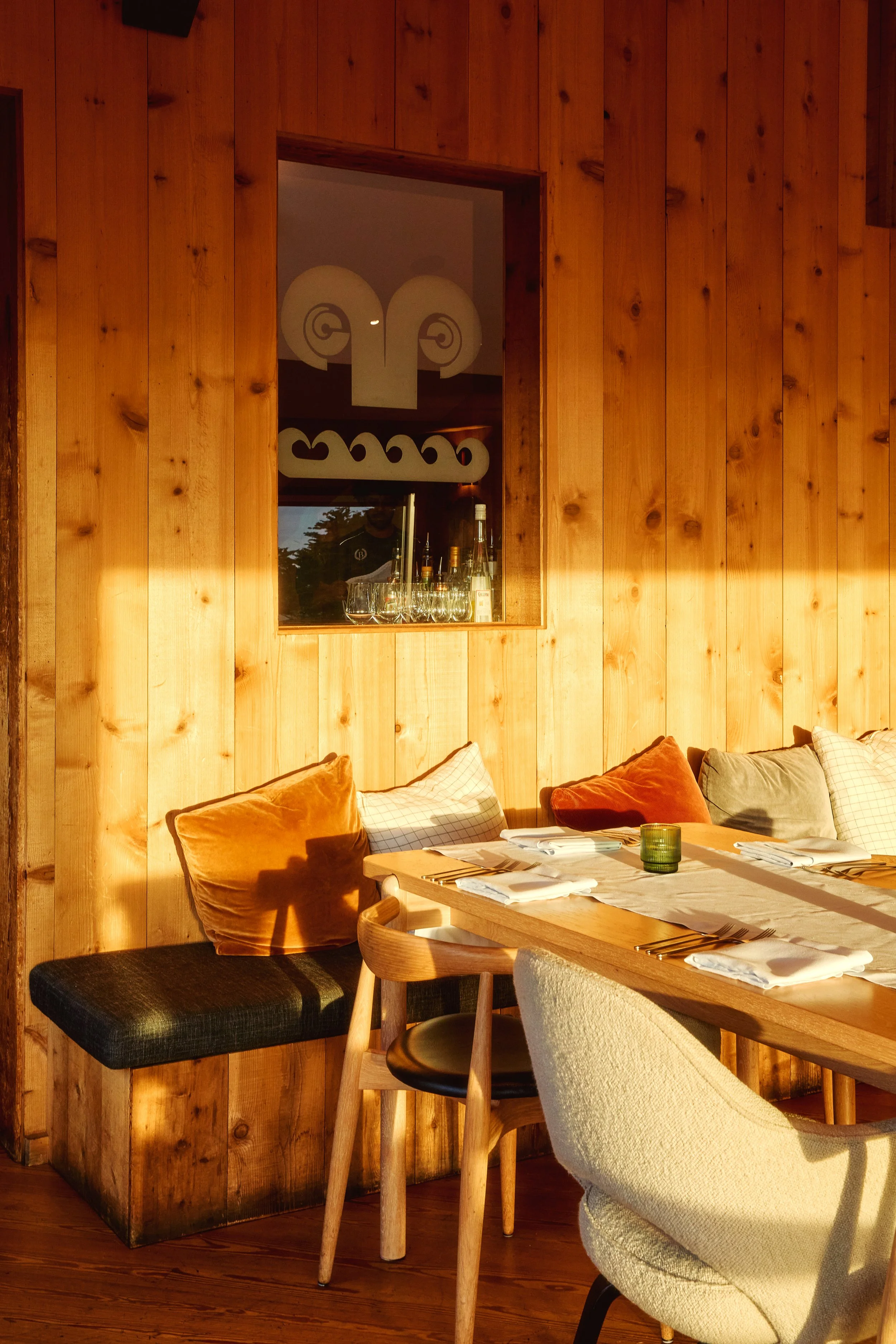 Sunlit dining area with a light wood table set with white napkins, silverware, and a green candle holder, surrounded by assorted chairs and cushions against a wooden paneled wall.