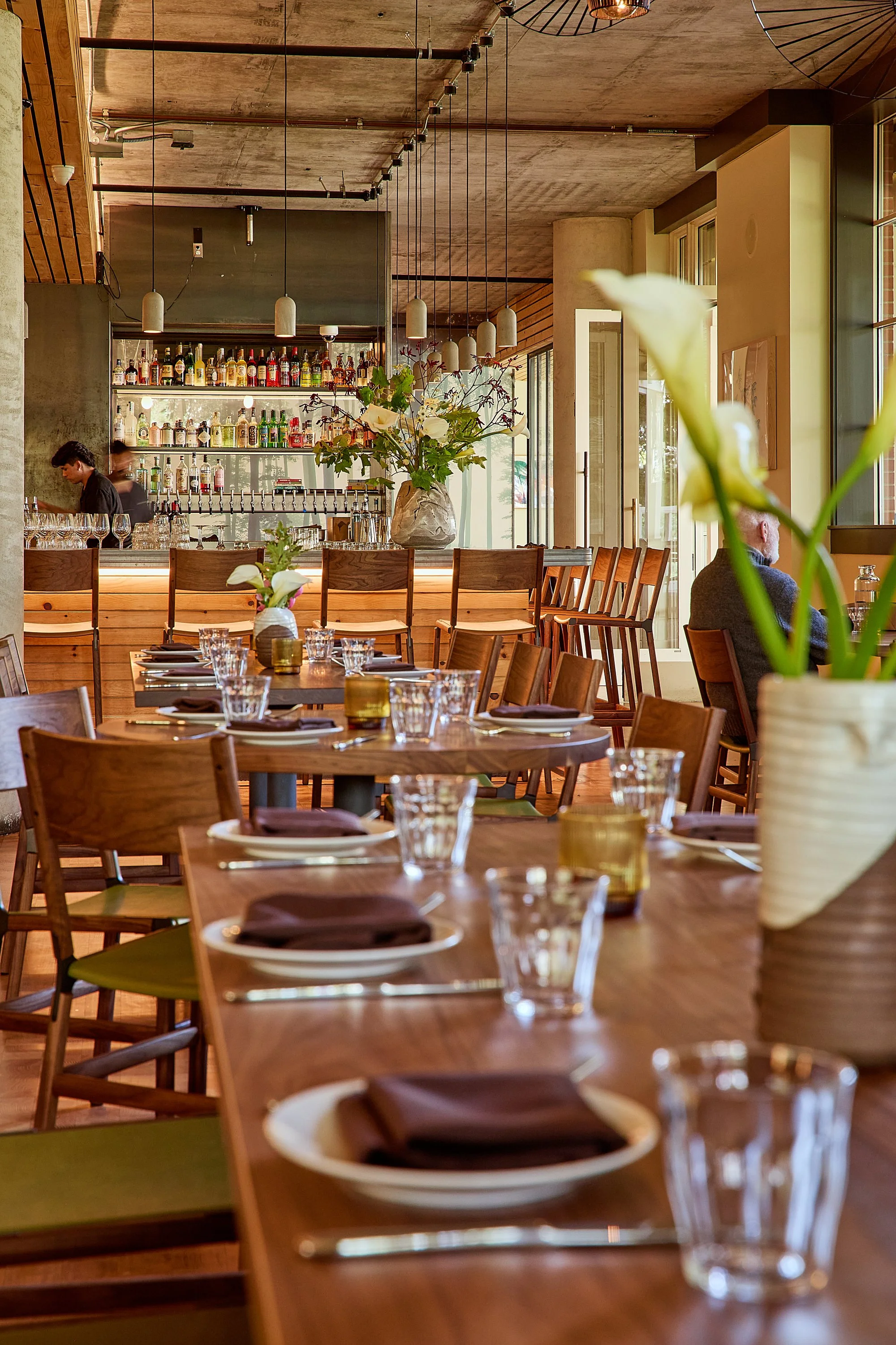 Interior of a restaurant or bar with tables set with glassware, plates, and napkins, a bar counter with bottles of alcohol, and large flower vases with flowers as decor.