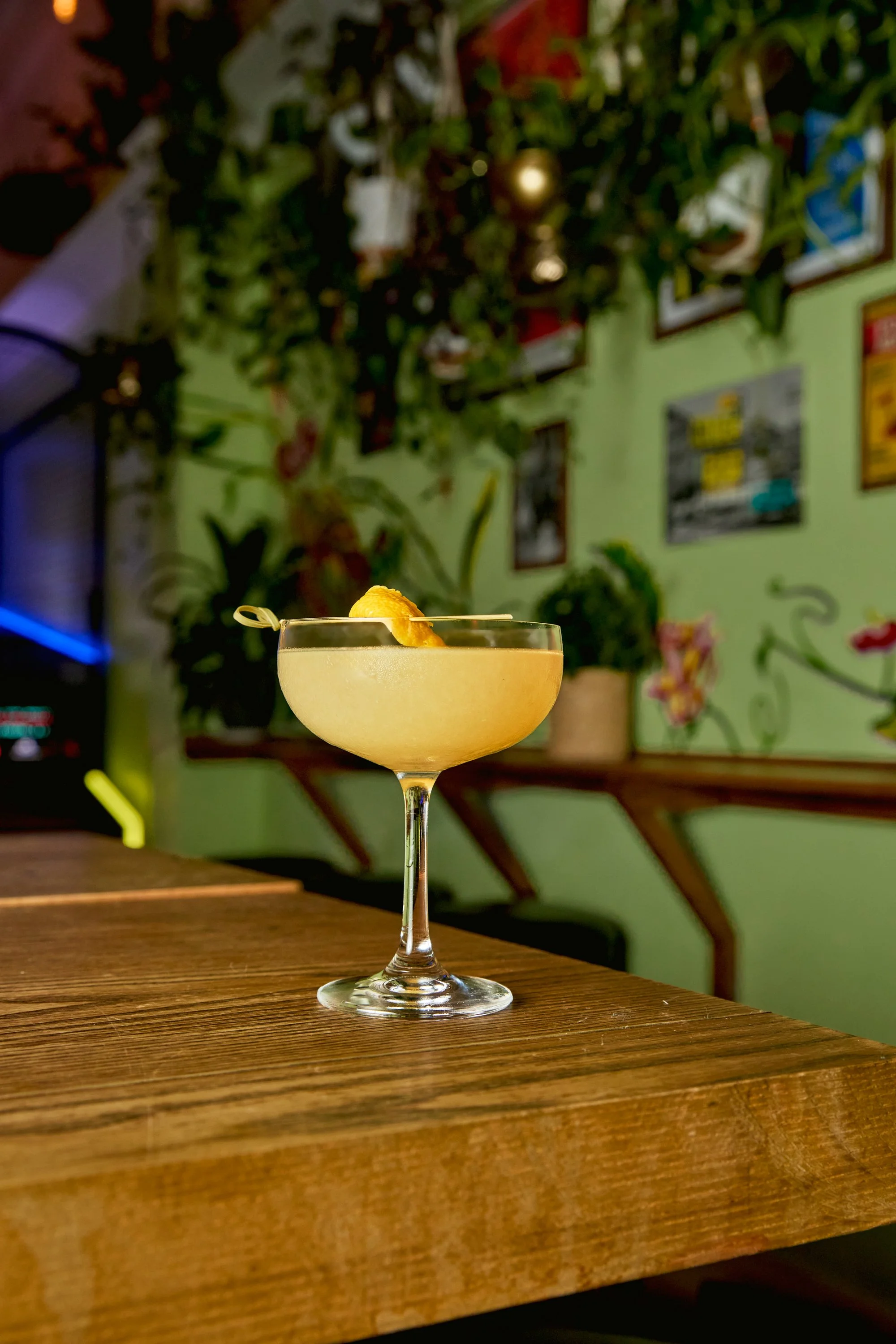 A yellow cocktail with a lemon twist in a coupe glass on a wooden bar counter in a dimly lit bar or restaurant with a green wall, framed pictures, and plants in the background.