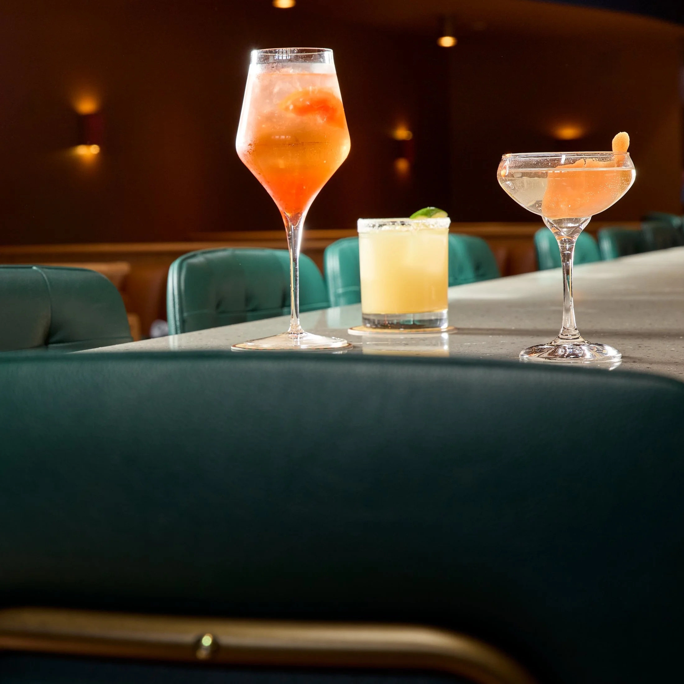 Two cocktails and a lime wedge on a bar table. The cocktail on the left is in a tall, curvy glass and contains a pinkish drink with ice and a cherry. The cocktail on the right is in a wide, shallow glass with a pinkish-orange drink, a gummy candy gar