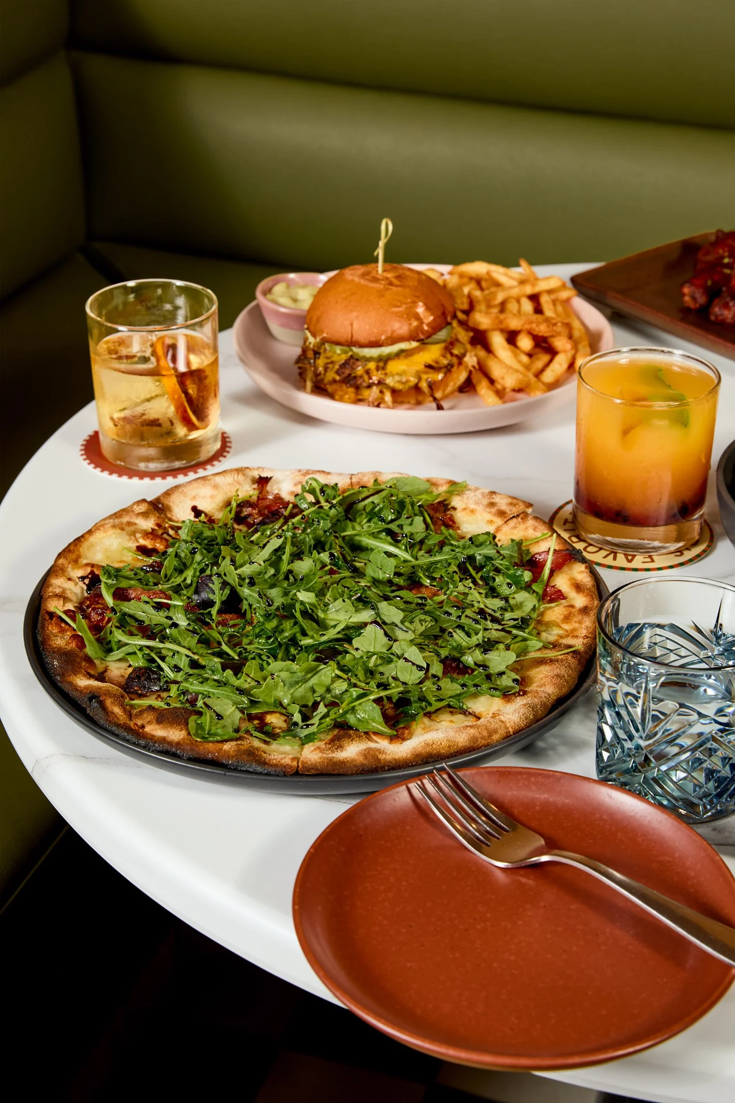 A pizza with arugula topping on a white table with a fork and a glass of water, a burger with fries and a drink, and a plate of chicken wings.