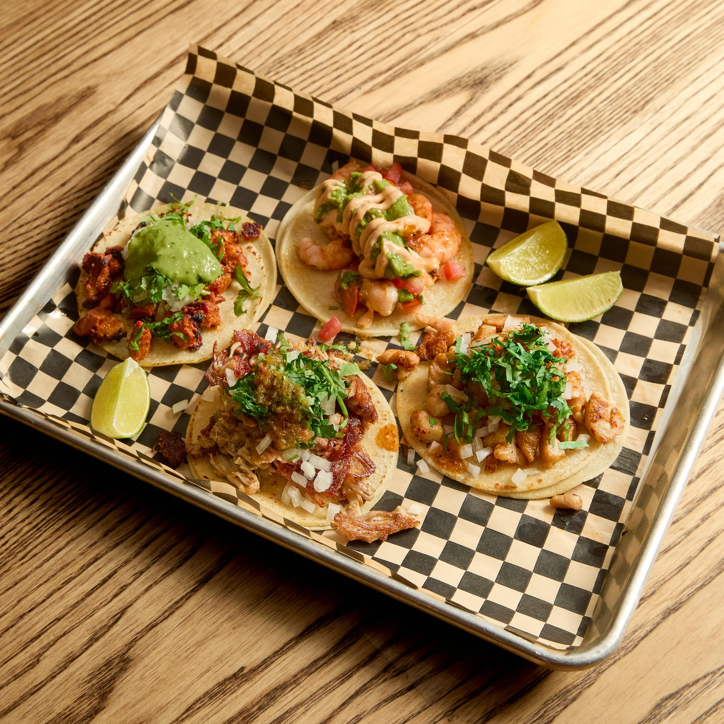 A tray with four tacos topped with various ingredients and three lime wedges on a checkered paper liner.