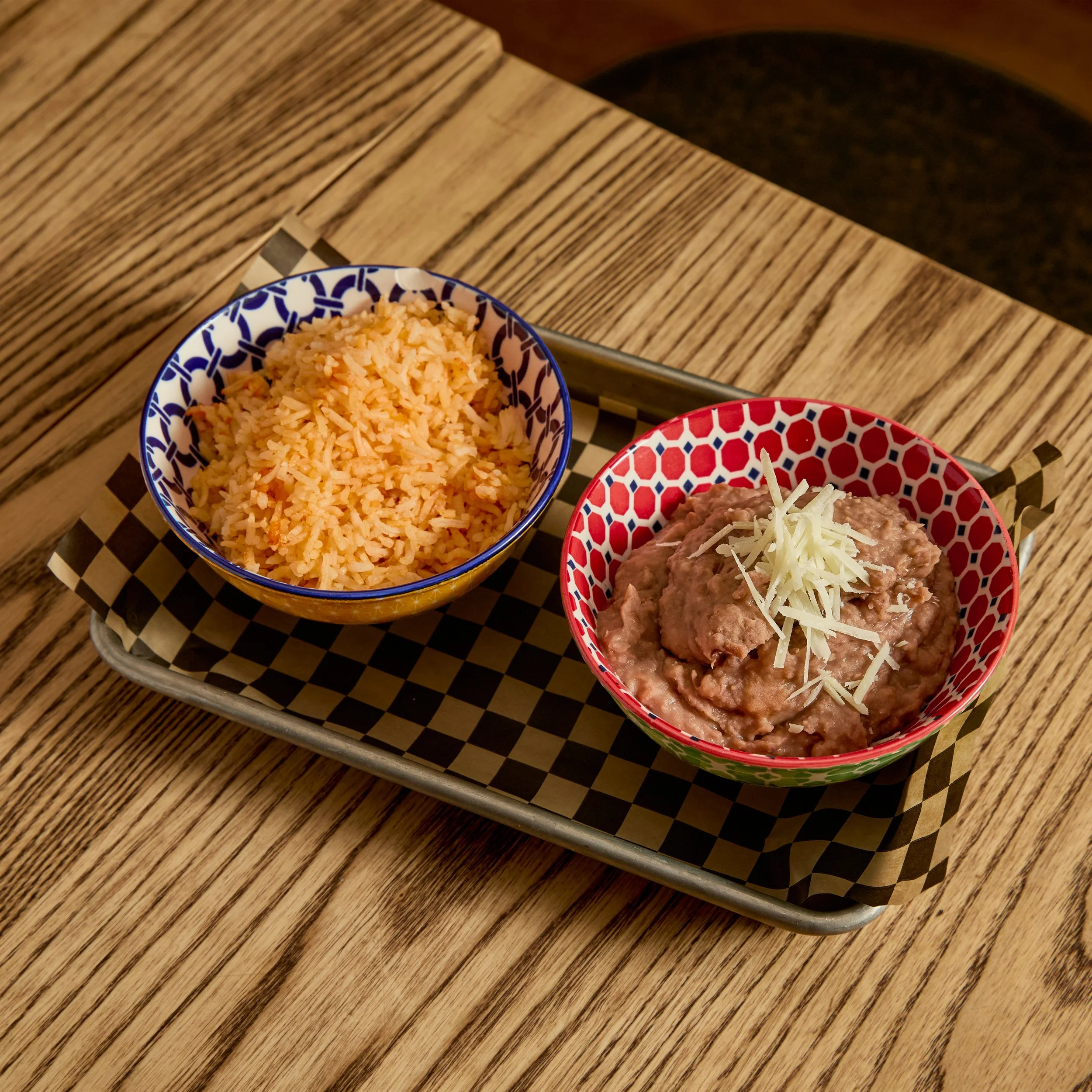 Bowls of rice and refried beans topped with shredded cheese on a checkered paper-lined tray on a wooden table.