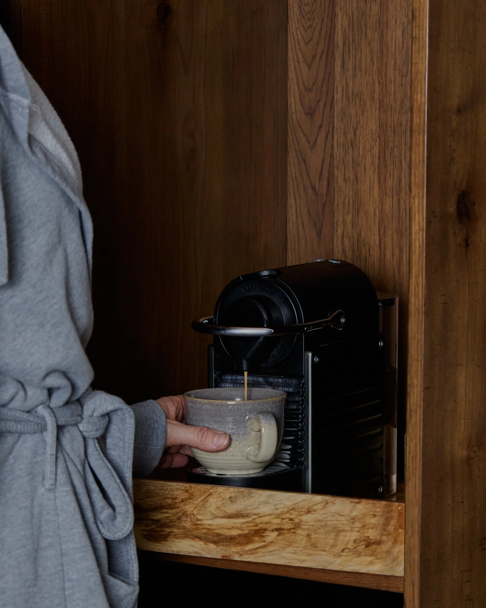 Person pouring coffee from a coffee machine into a mug inside a wooden kitchen cabinet.