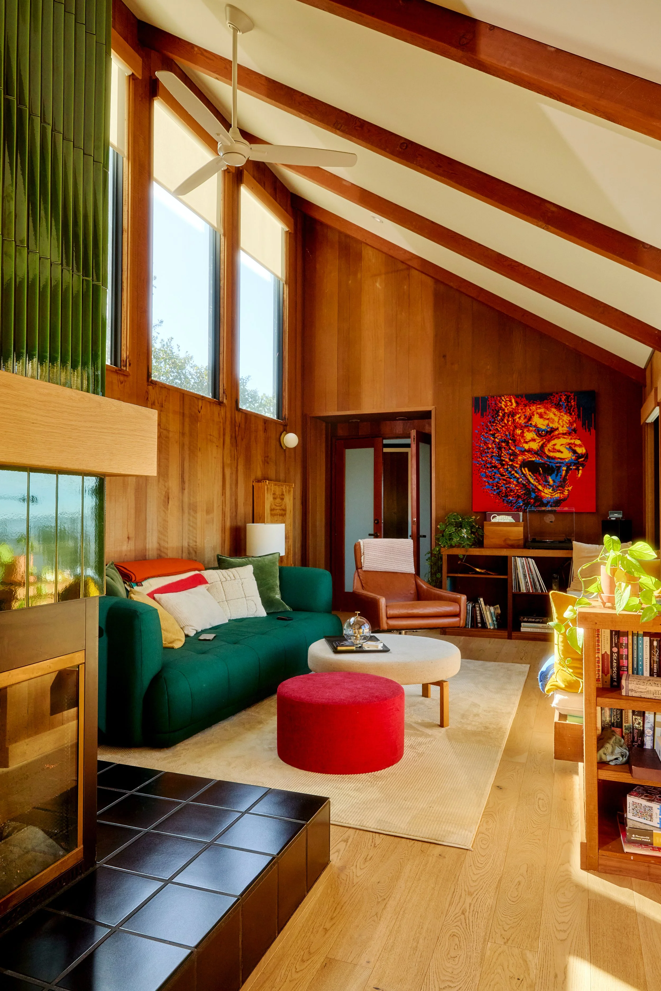 Living room with wooden walls and ceiling beams, large windows, green couch with multiple cushions, round red ottoman, beige rug, white round coffee table with glass globe and remote, leather armchair, bookshelves, colorful lion artwork on wall, pott