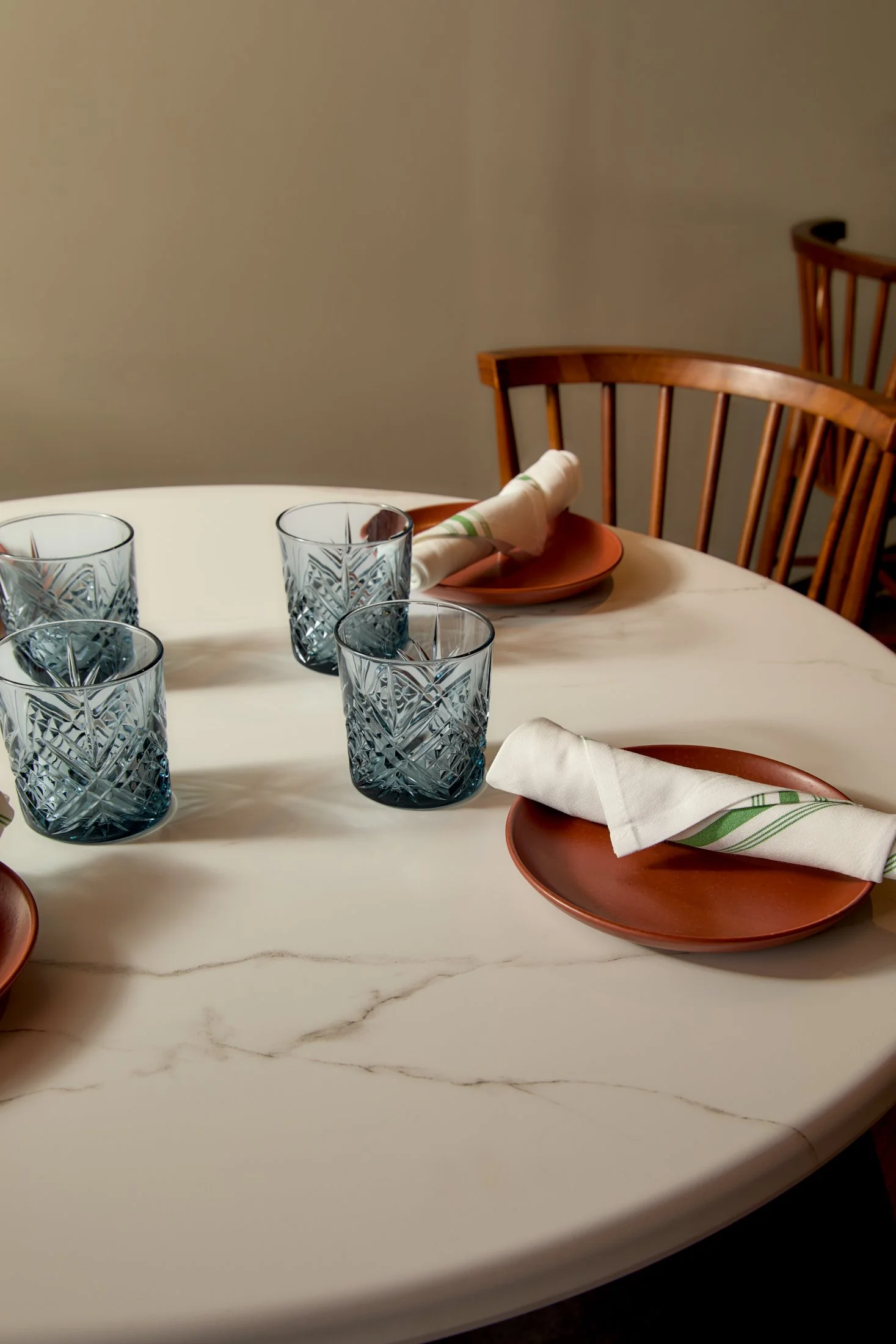 A dining table set with four textured glass cups, two orange plates with white napkins wrapped in green-striped cloth, and wooden chairs against a beige wall.