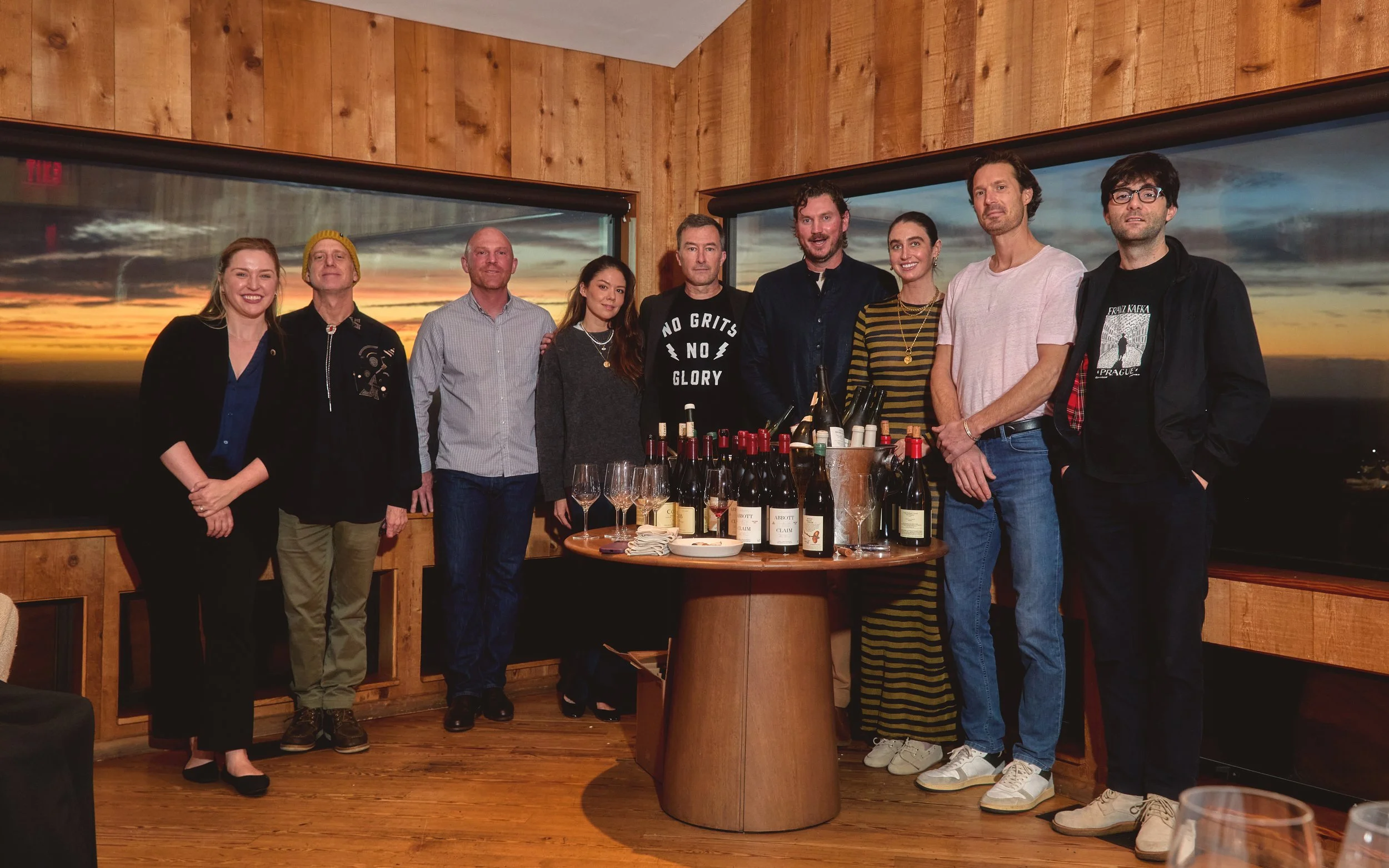 Group of ten people standing behind a round table with wine bottles, glasses, and other items, inside a room with wooden walls and large windows showing a sunset sky.