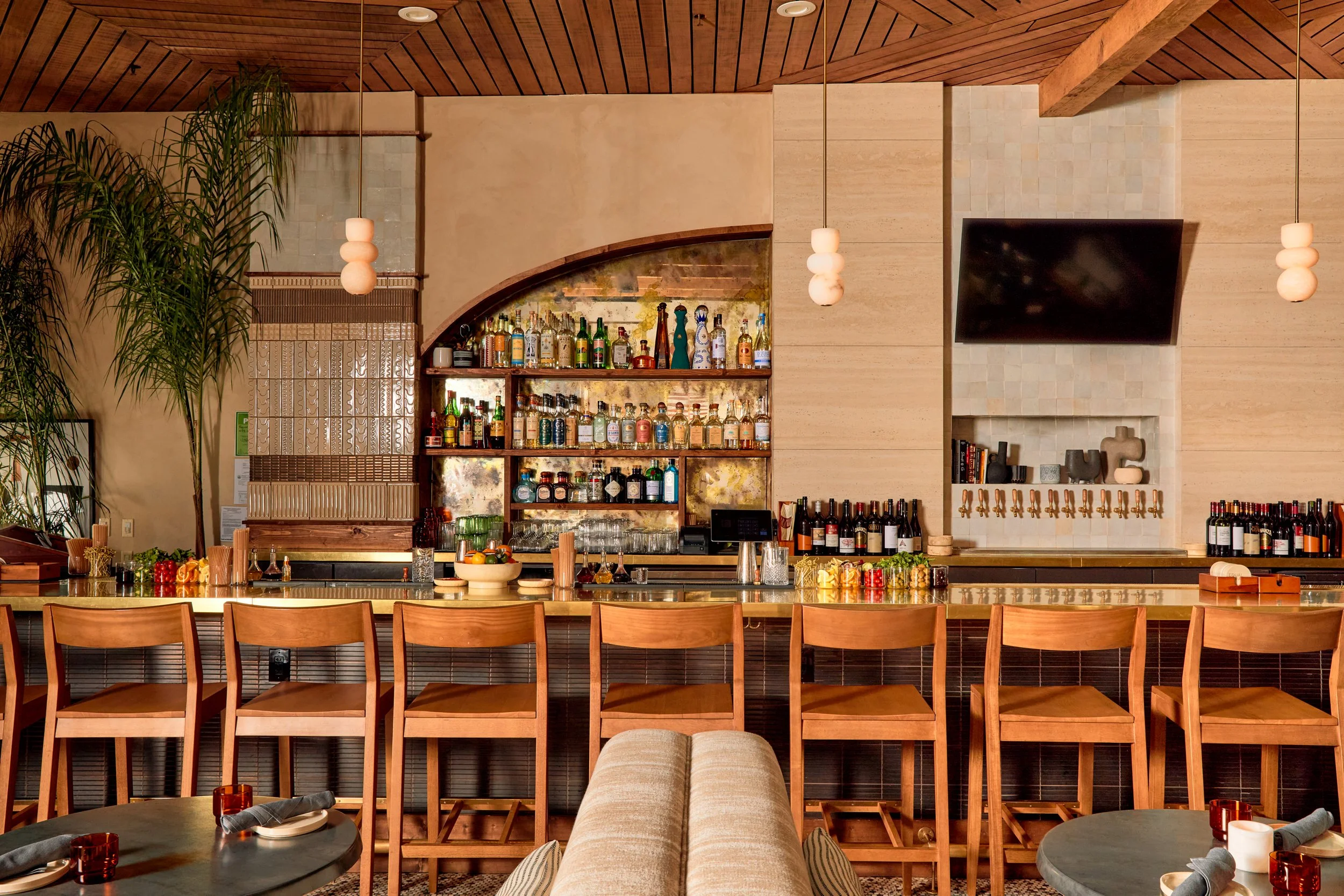Interior of a restaurant or bar with a long wooden bar counter, chairs, bottles of alcohol on shelves, a TV, and modern decor including hanging lights and greenery.