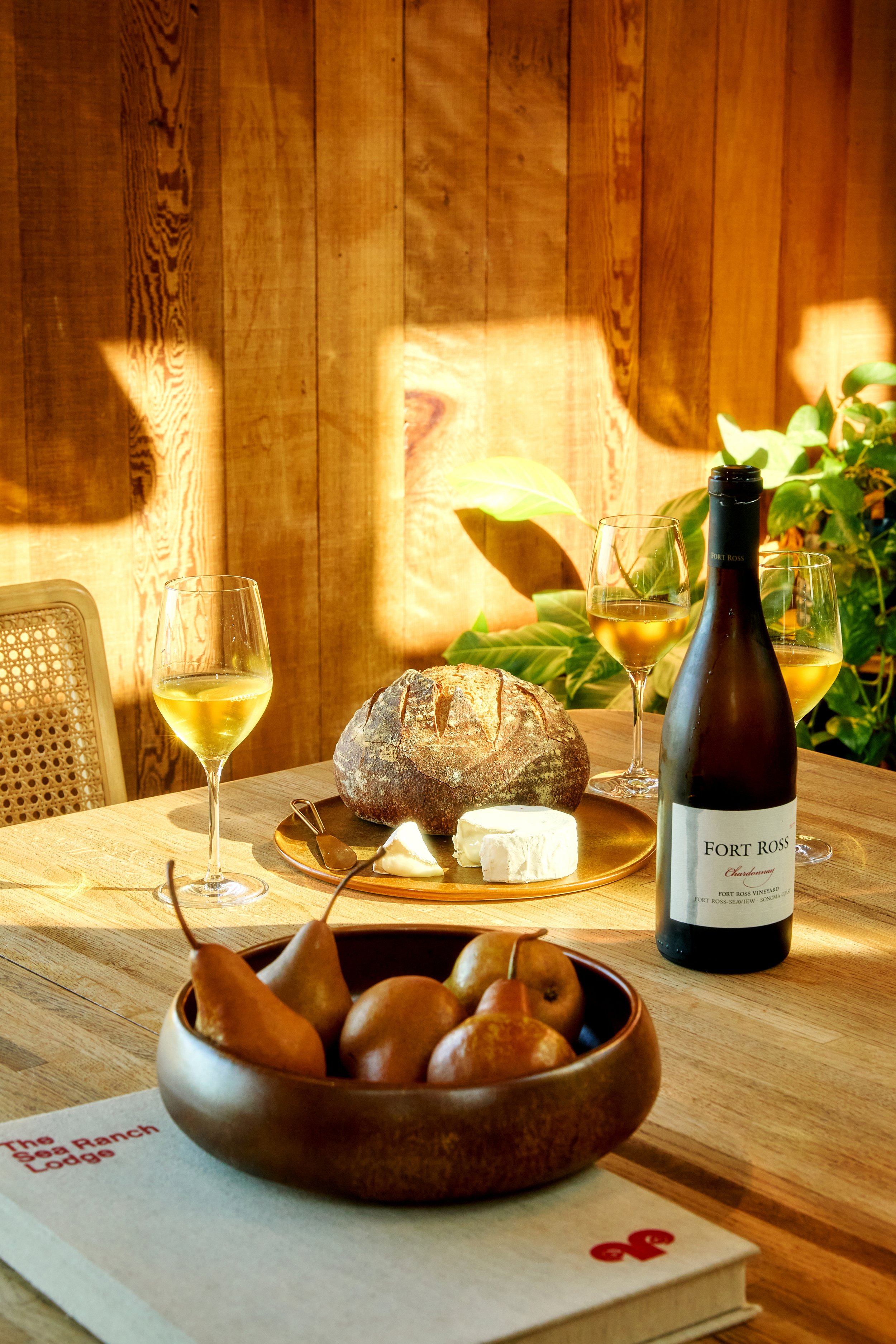 A wooden table set with a loaf of bread, cheese, three glasses of white wine, a bottle of Fort Ross Chardonnay, a bowl of pears, and a book on a wooden wall background with sunlight.