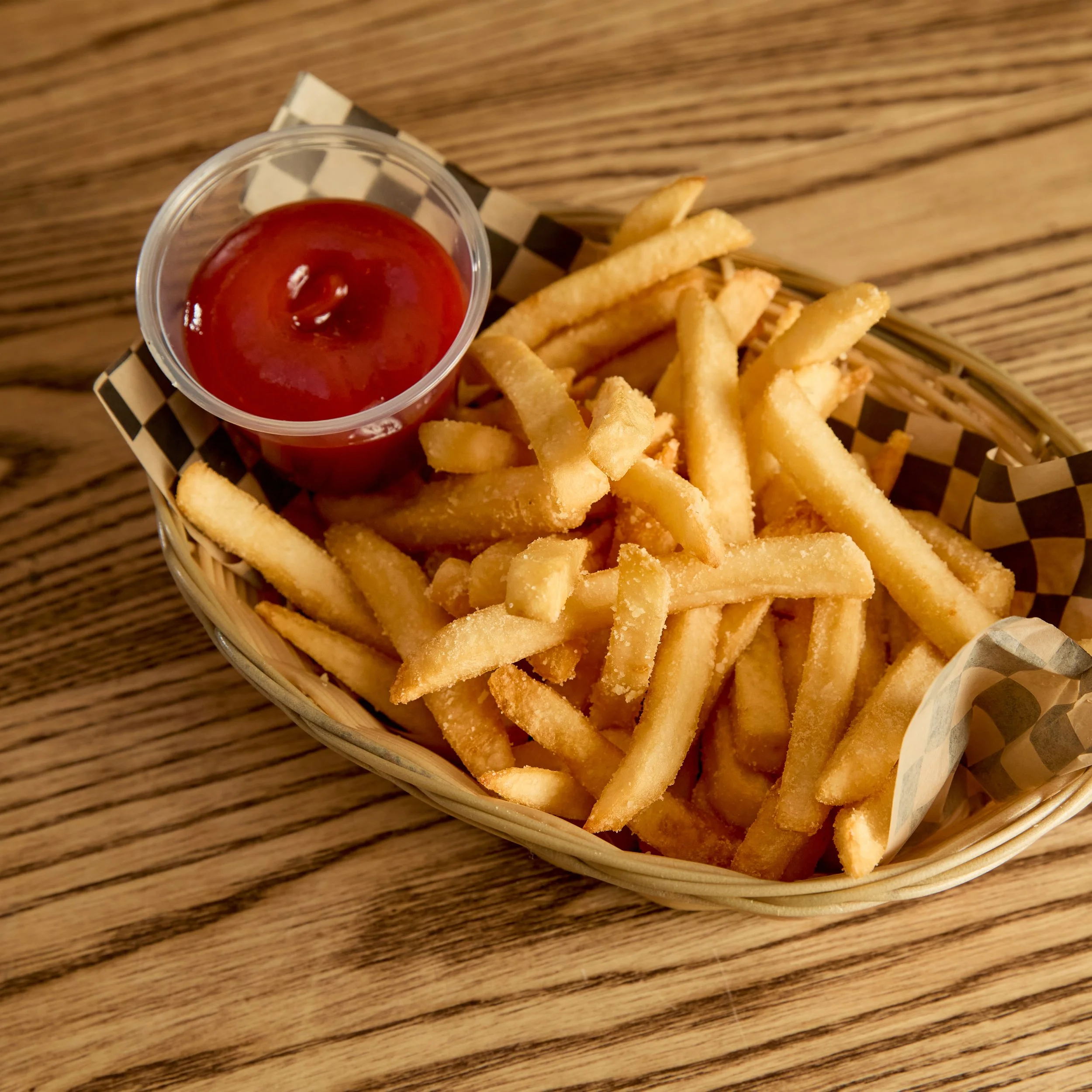 Basket of French fries with ketchup on a checkered paper liner on a wooden table.
