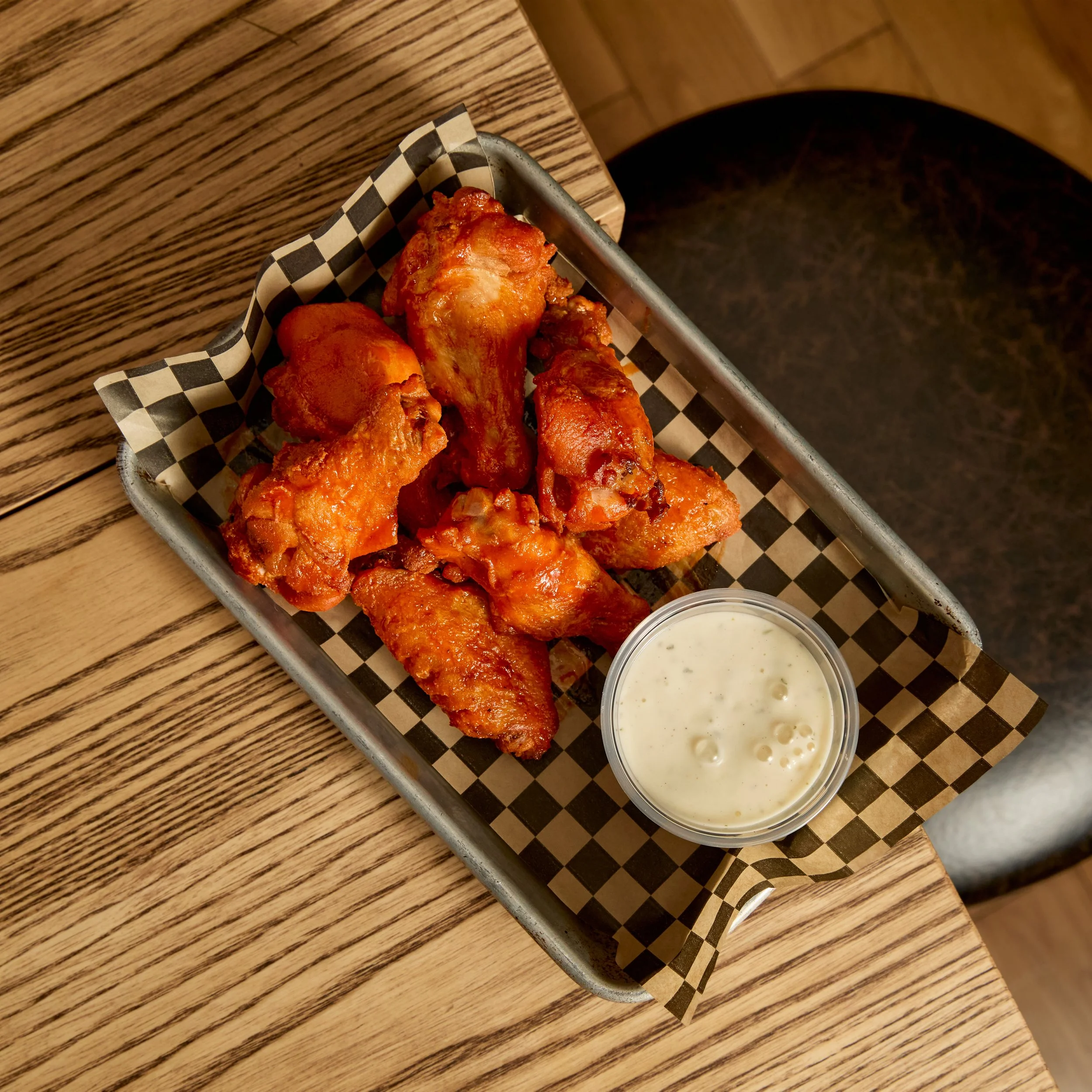 Bowl of buffalo chicken wings with a side of ranch dipping sauce on a checkered paper liner in a metal basket on a wooden table.