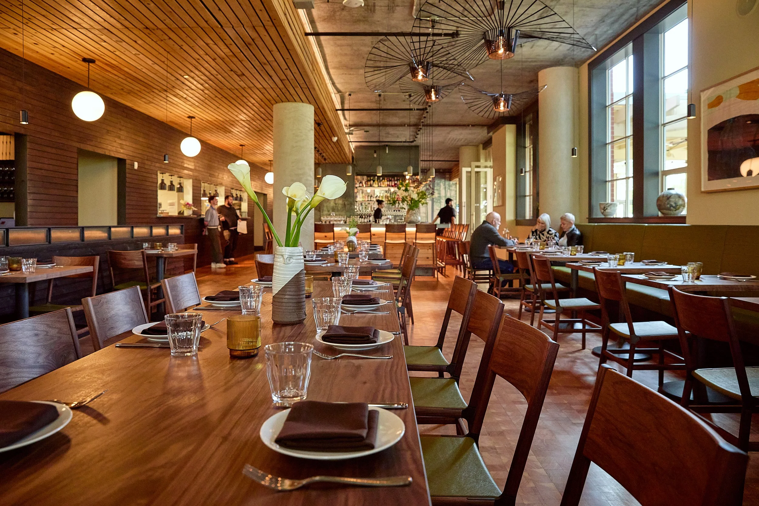 Interior of a modern restaurant with wooden tables and chairs. A vase with white calla lilies is on the table in the foreground. There are people dining and staff working, with large windows letting in natural light. Stylish ceiling lights and art on