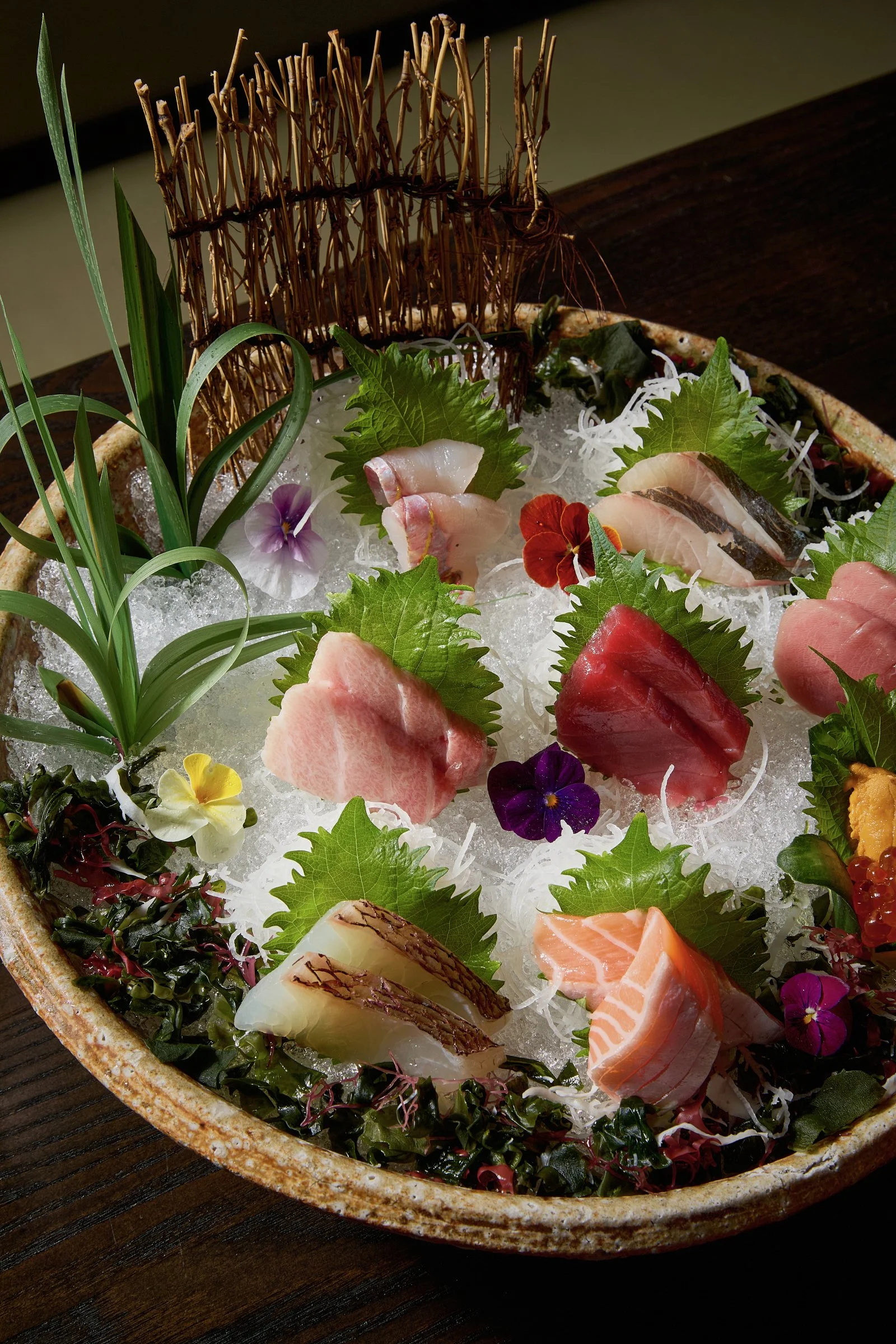 An assortment of sashimi slices with edible flowers and green leaves on a bed of ice in a ceramic dish.