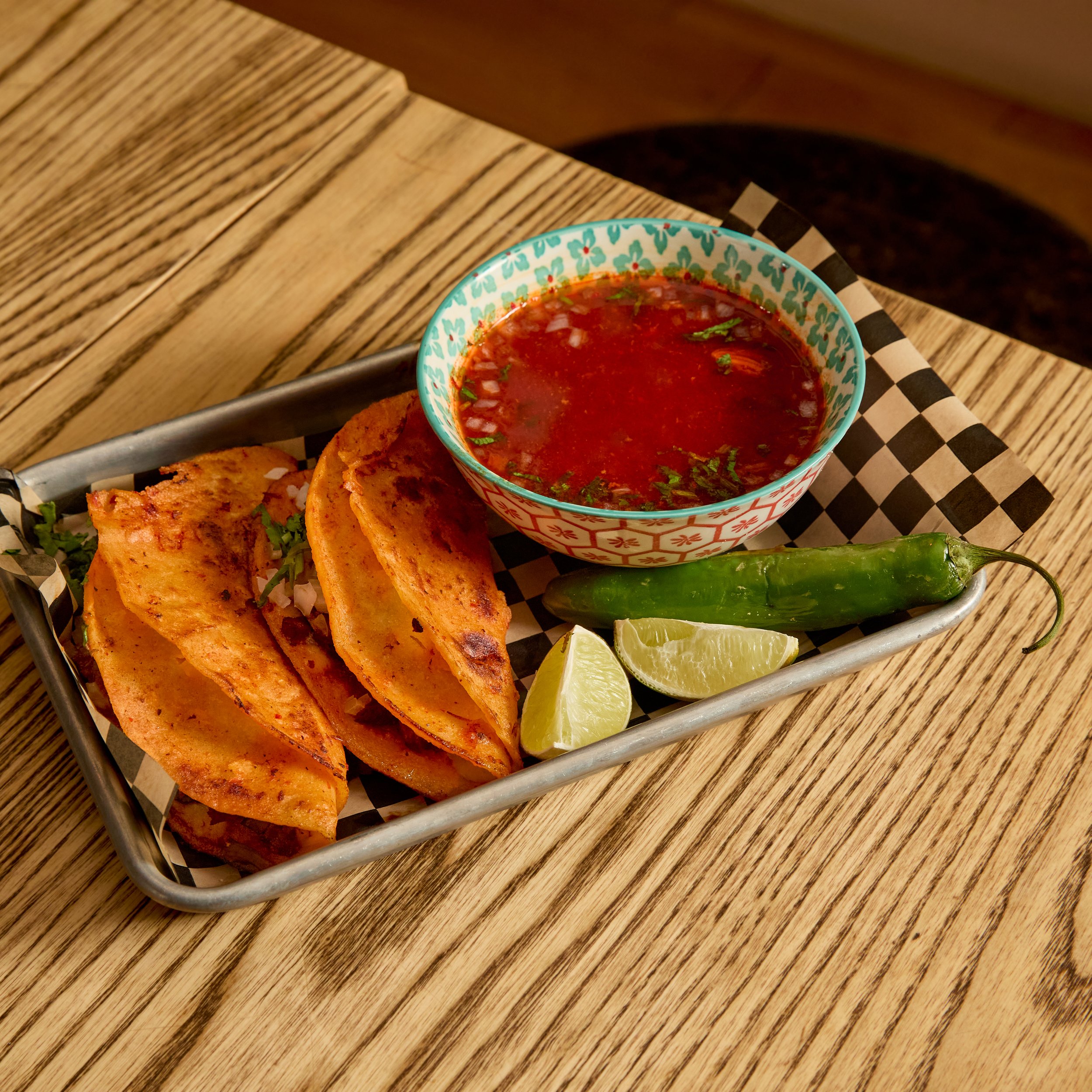Fried tacos, lime wedges, a jalapeño pepper, and a bowl of red salsa on a metal tray with black and white checkered paper, on a wooden table.