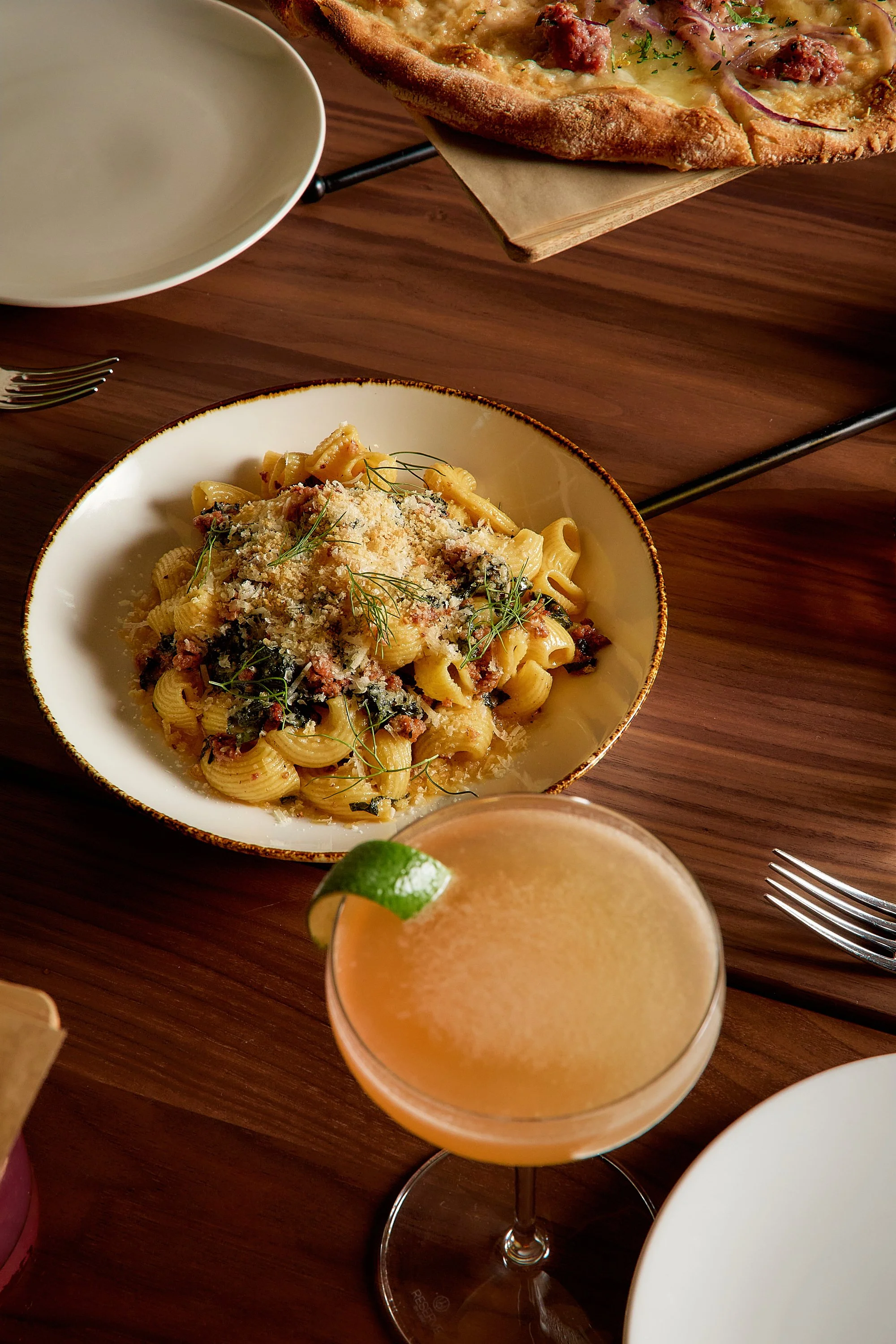 A close-up of a plate with pasta topped with grated cheese and garnished with herbs, a cocktail with a lime wedge, and a pizza in the background on a wooden table.
