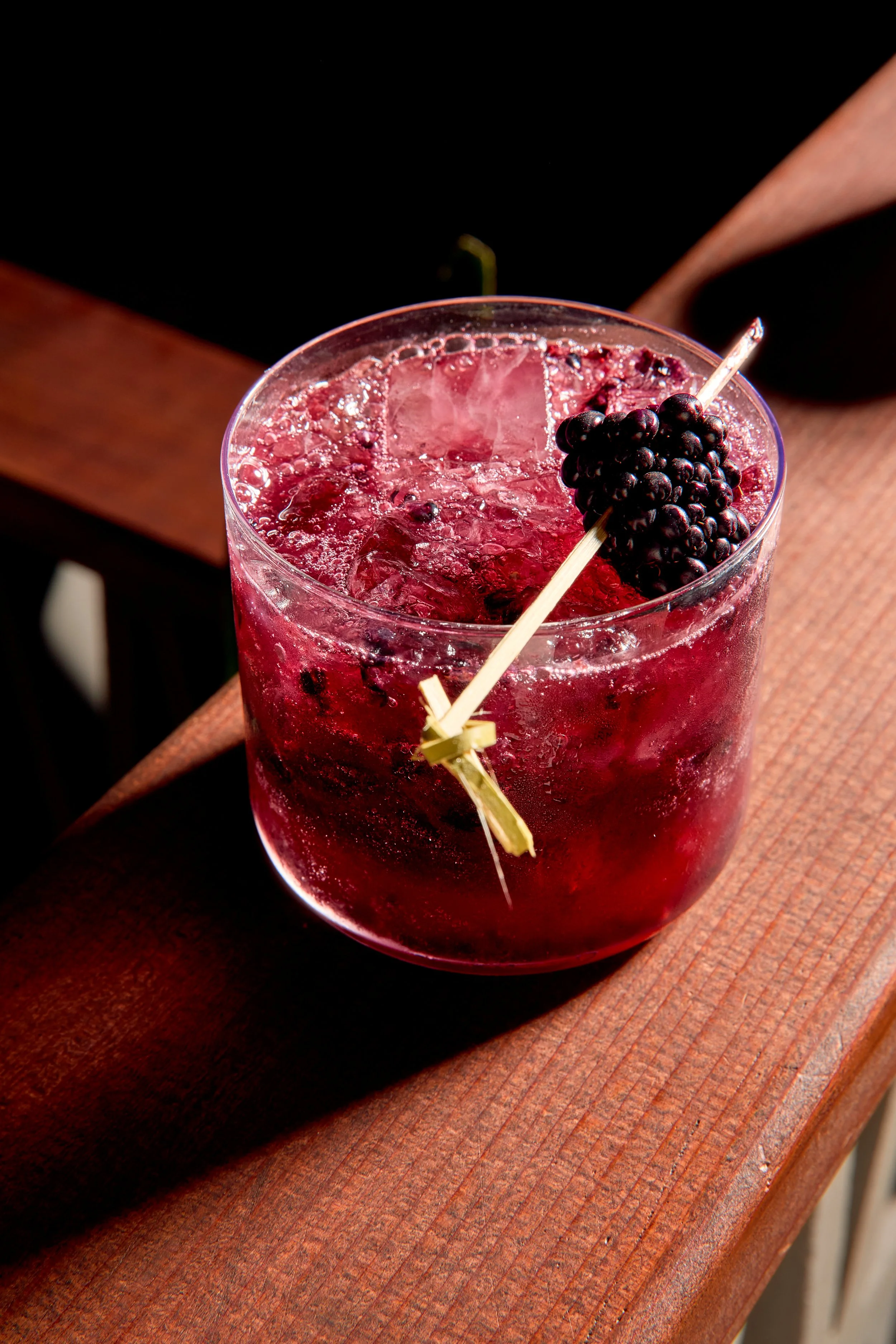 A tall glass of a dark red berry cocktail with ice cubes, garnished with a cluster of blackberries and a decorative bamboo skewer resting across the glass, placed on a wooden surface.