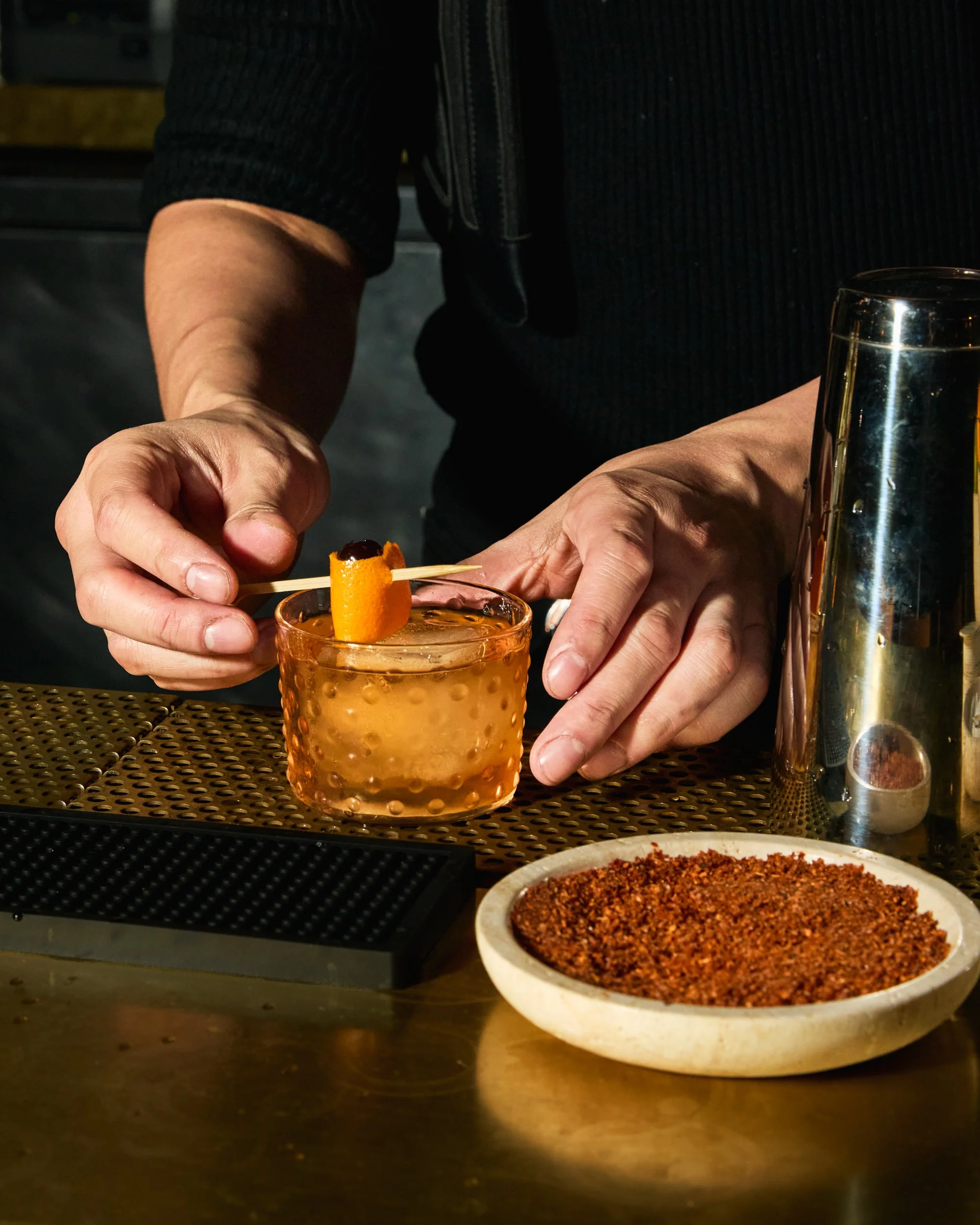 Bartender garnishing an orange twist in a cocktail at a bar with a bowl of spice on the table.