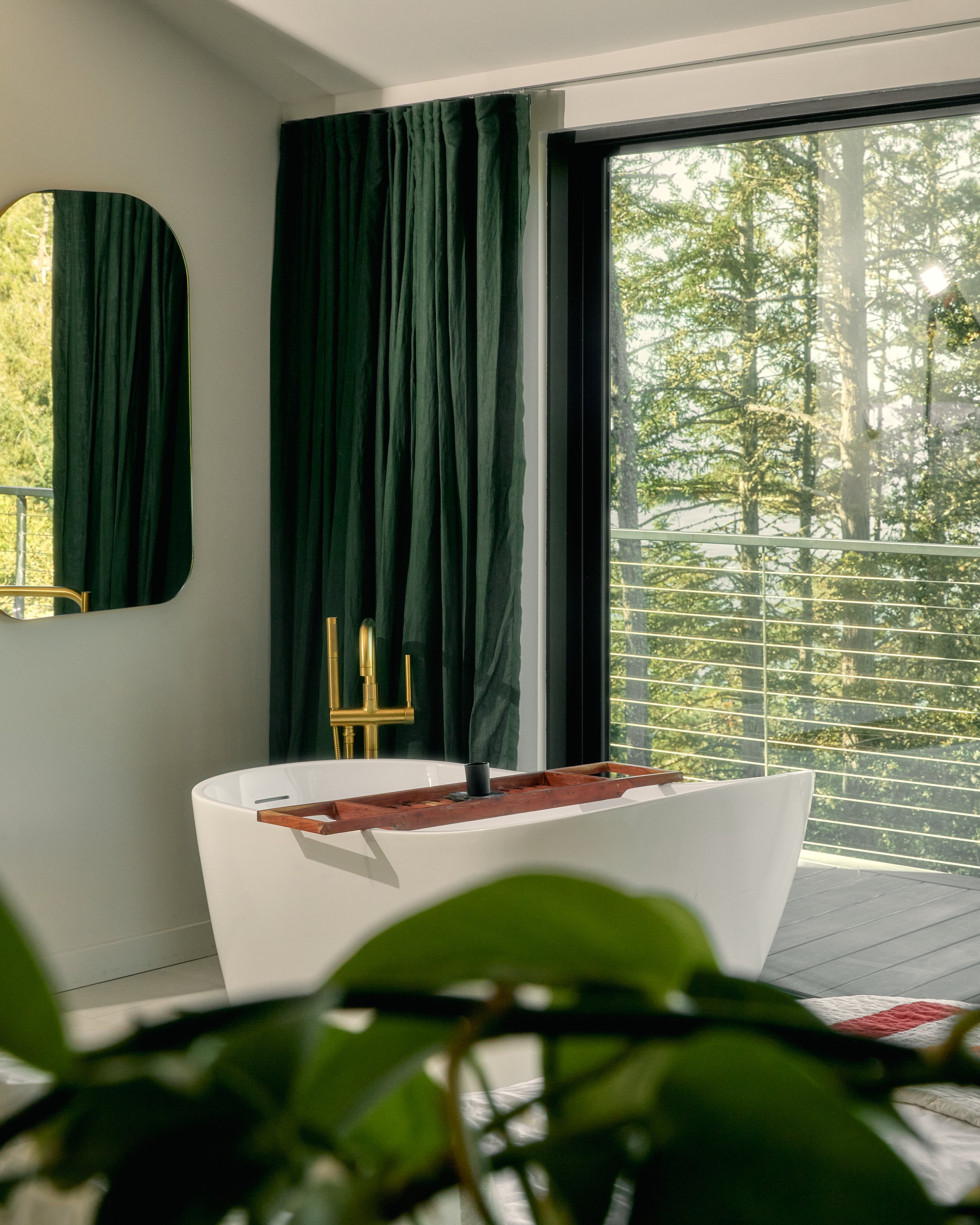 Modern bathroom with a white freestanding bathtub, green curtains, a large window overlooking trees, a gold faucet, and a mirror reflecting the outdoor scenery.
