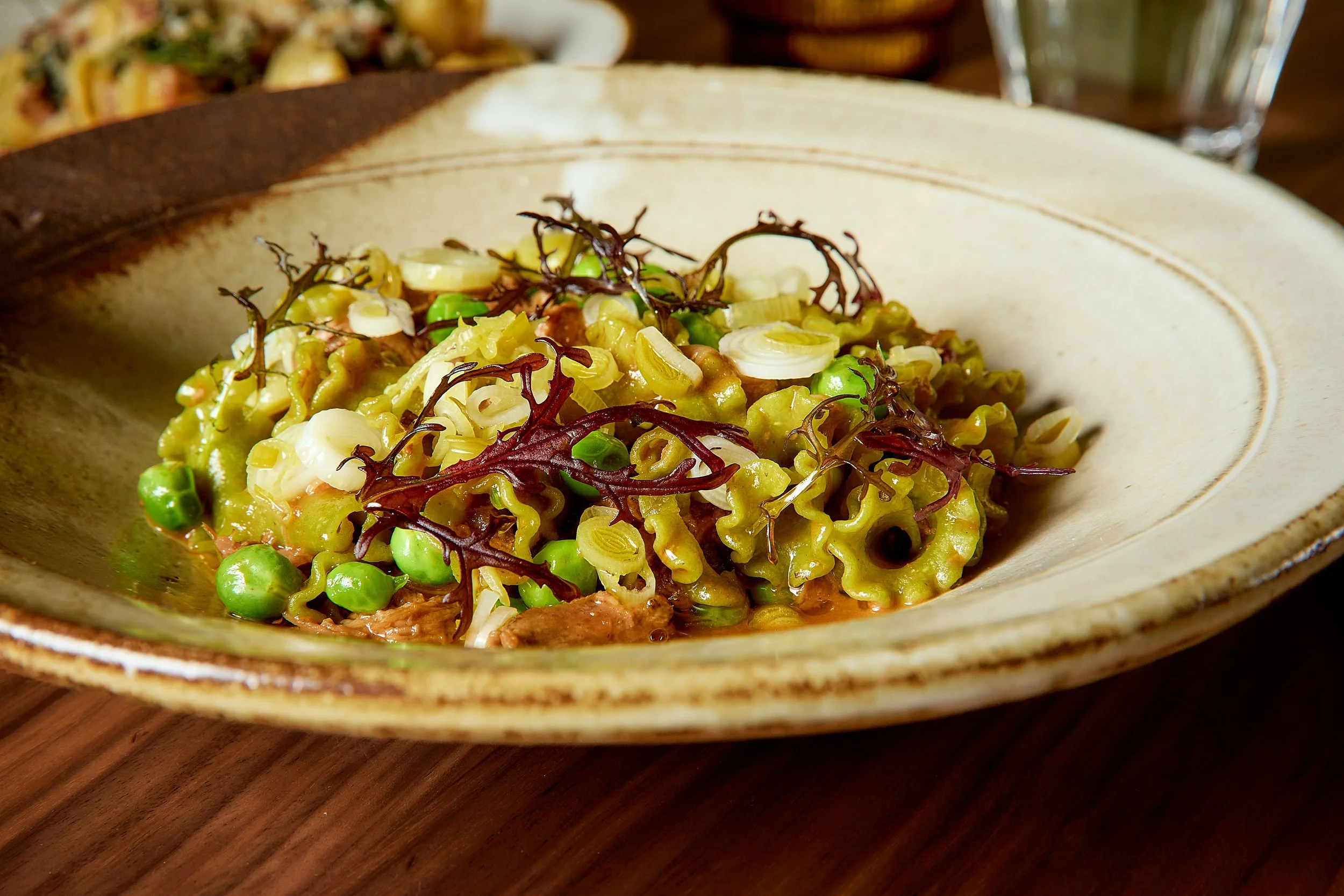 Bowl of pasta with green peas, thinly sliced leeks, and garnished with red seaweed, on a wooden table.