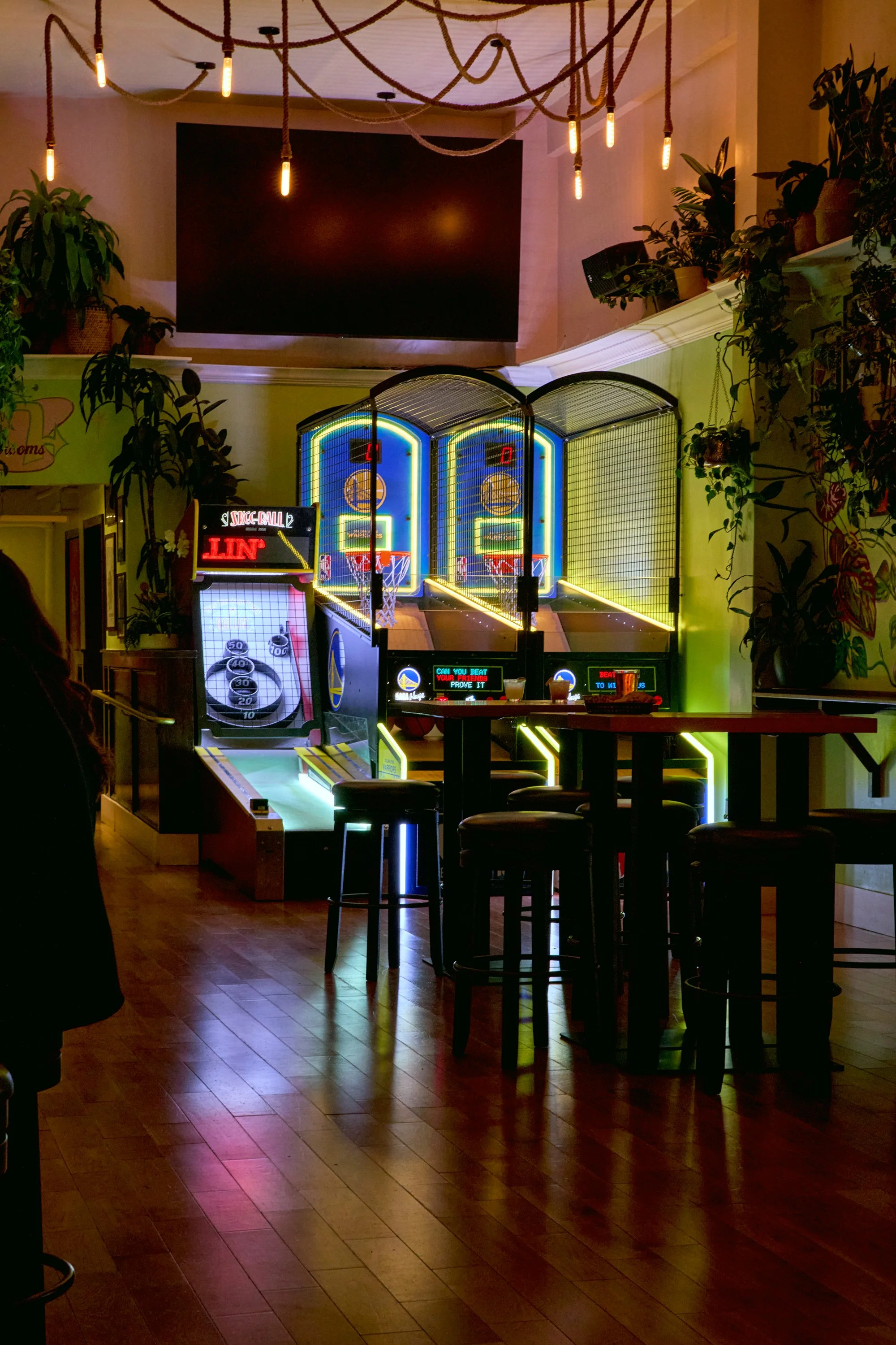 Indoor arcade with a basketball shooting game and seating area, decorated with plants and string lights.
