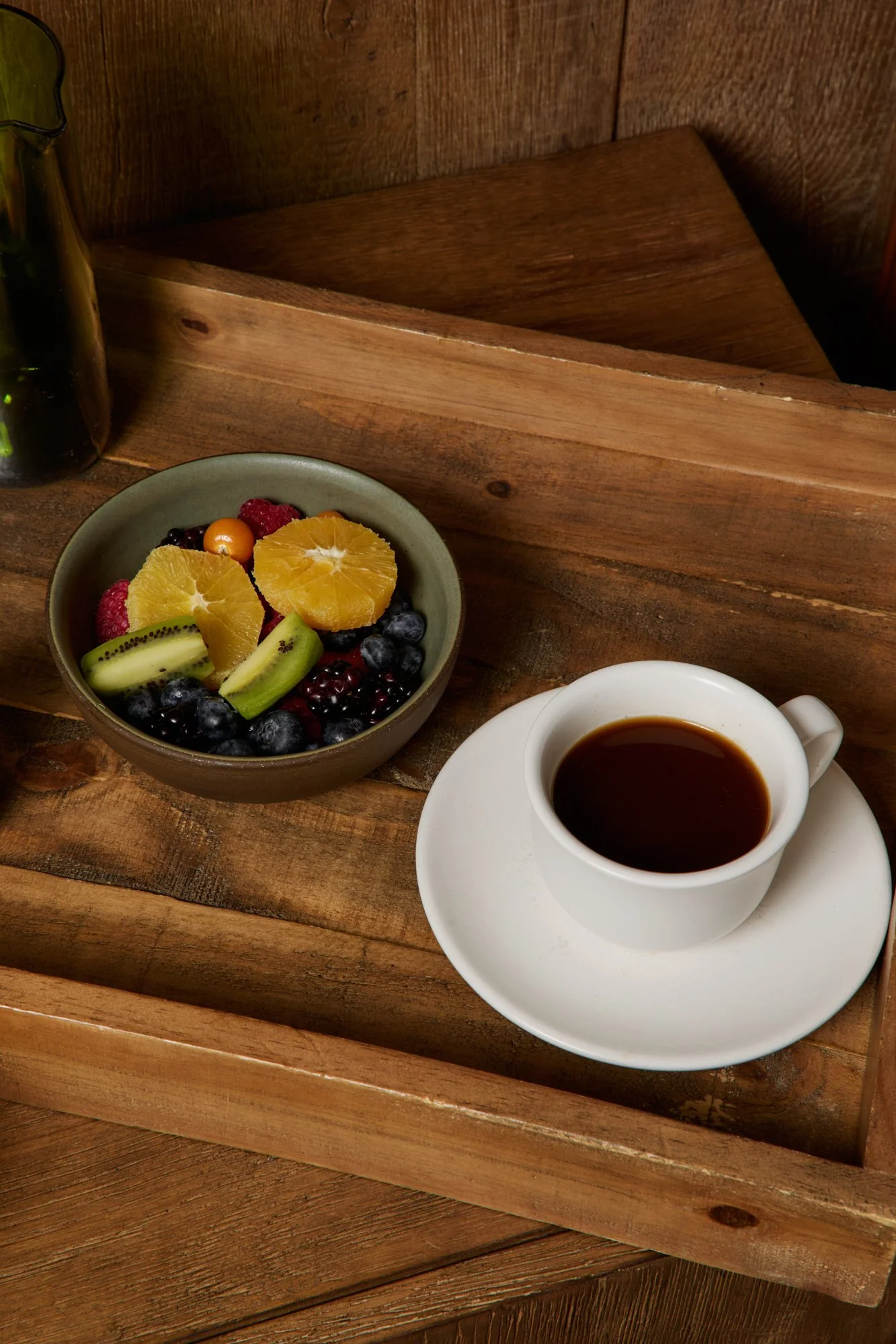 A wooden tray with a cup of black coffee on a saucer, and a bowl of mixed fresh fruit including orange slices, kiwi, blueberries, raspberries, blackberries, and a small yellow cherry tomato. A dark glass bottle with a plant is partially visible on th