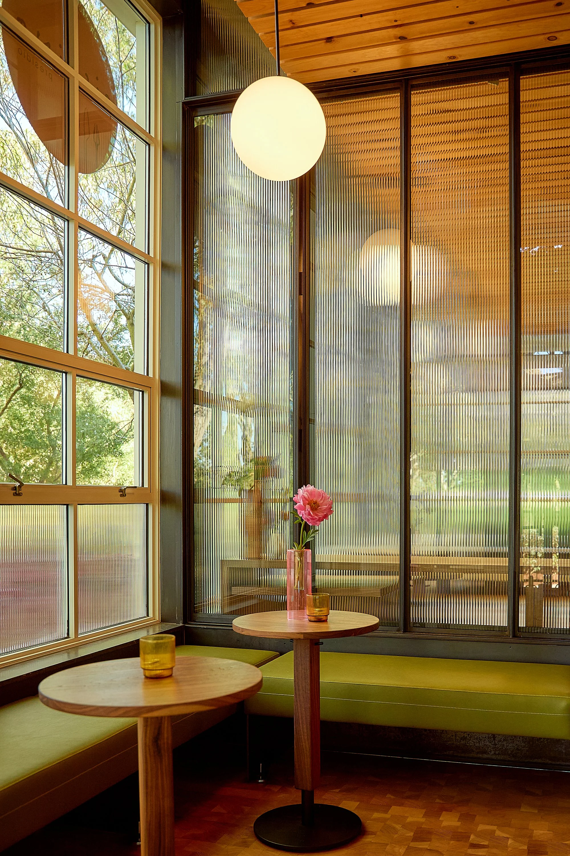 Interior of a room with large windows, wooden ceiling, and a wall made of vertical wooden slats. Small round tables with vases holding pink flowers and yellow cups are visible.