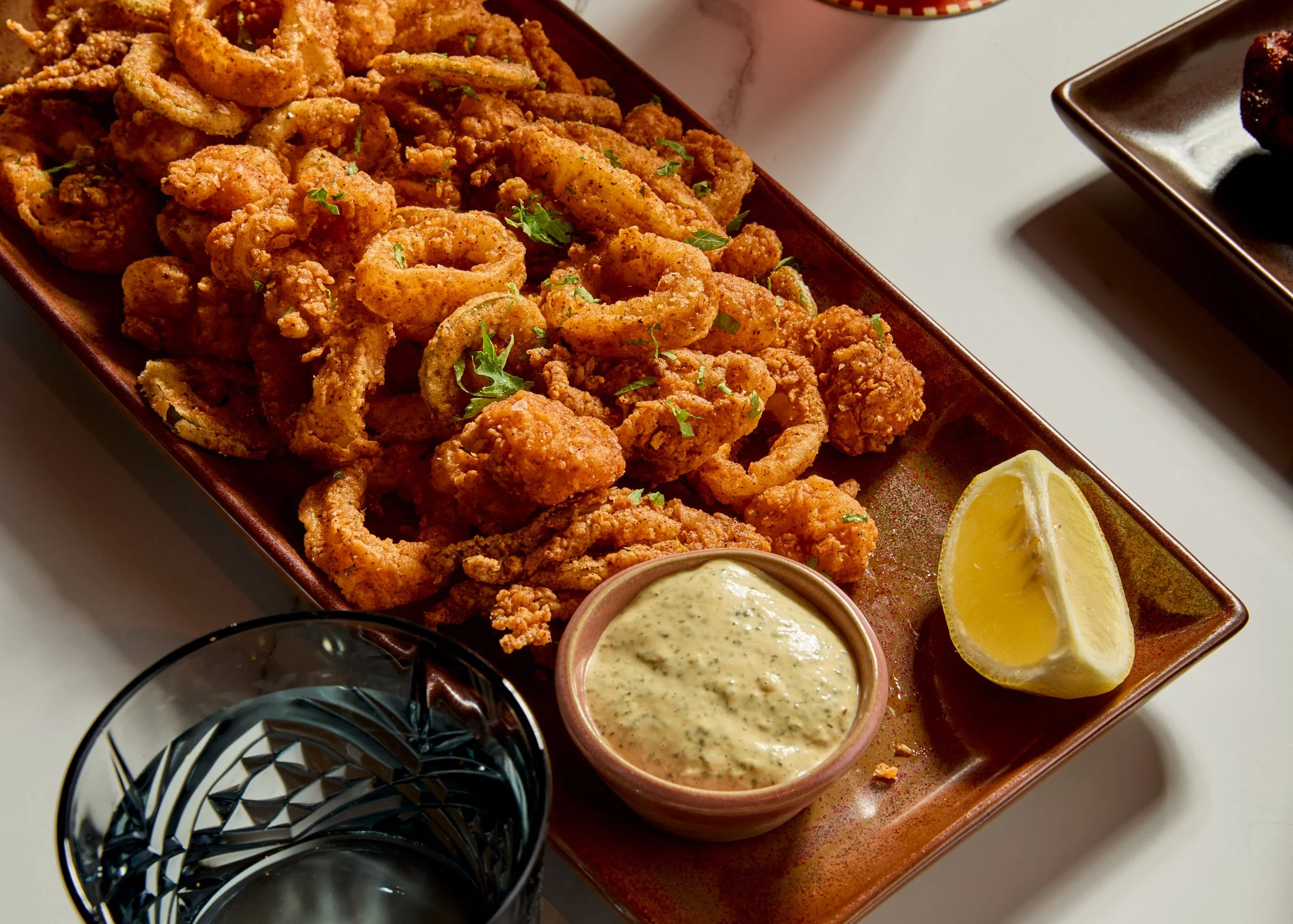 A tray of fried calamari with lemon wedges, creamy dipping sauce, and dark soy sauce on a white table.