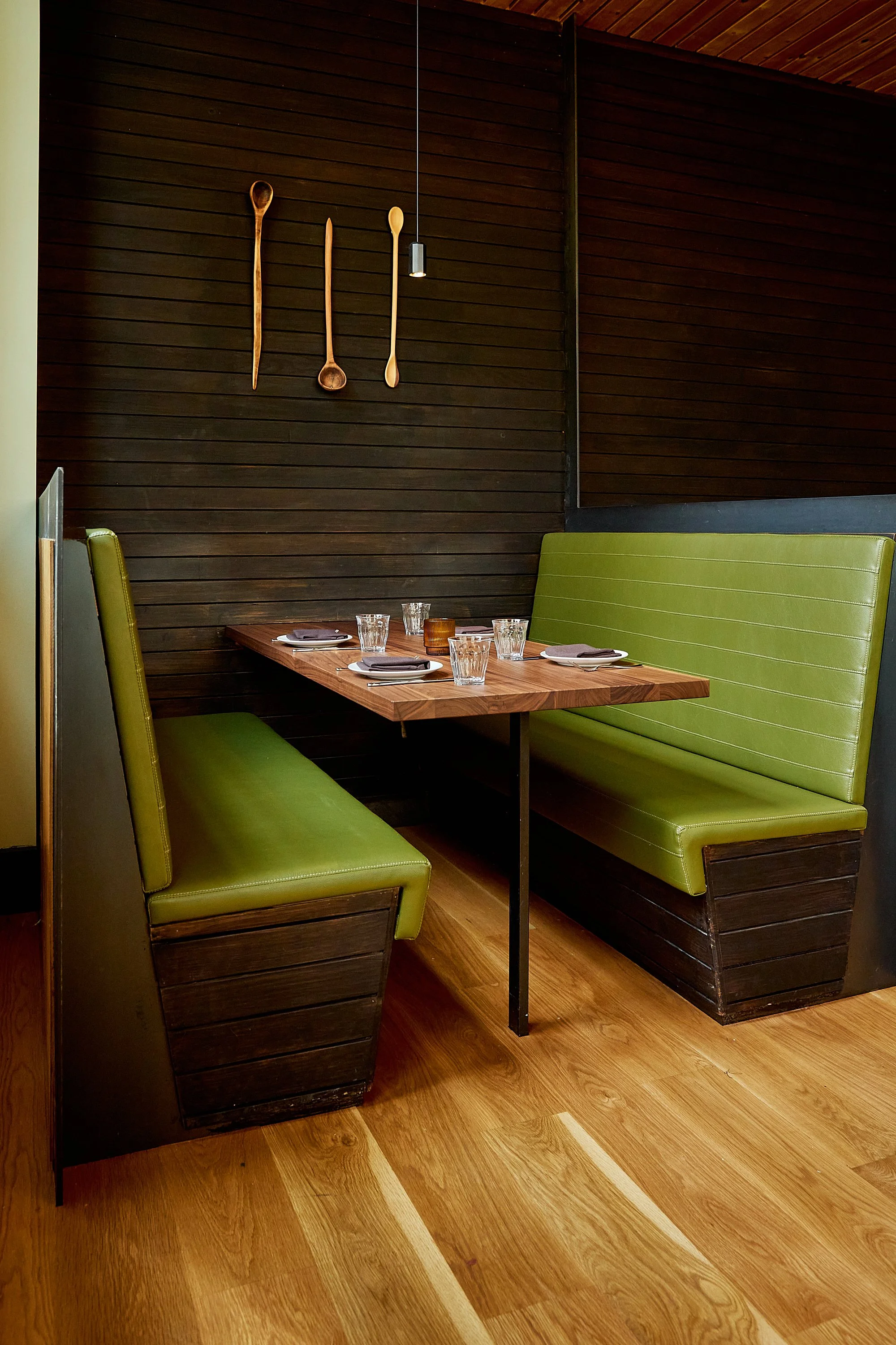A wooden dining table set for four with glasses, plates, and napkins in a cozy restaurant corner. Green upholstered booth seating surrounds the table, and a dark wooden wall with three wooden spoons and a letter opener hanging decorates the backgroun
