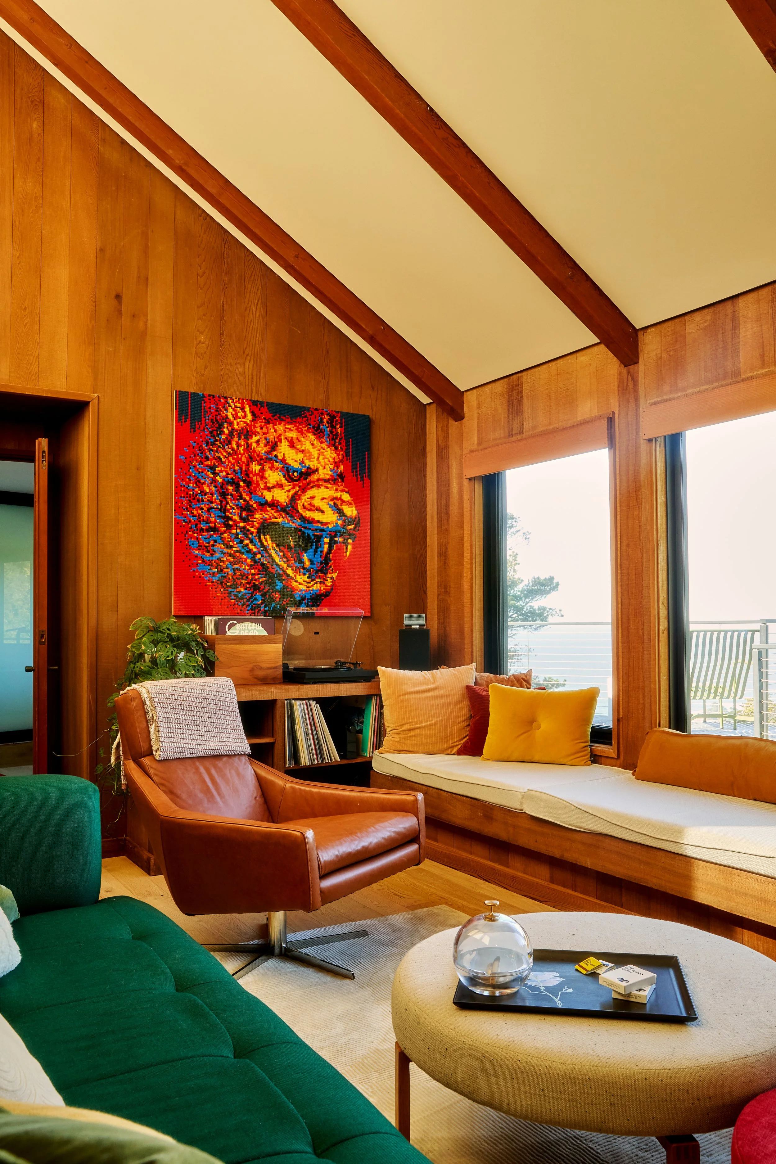 Living room with wood-paneled walls, a large window with an outdoor view, a colorful pixel art eagle on the wall, a brown leather armchair, a green sofa, and a round beige ottoman with a tray and small decorative items.