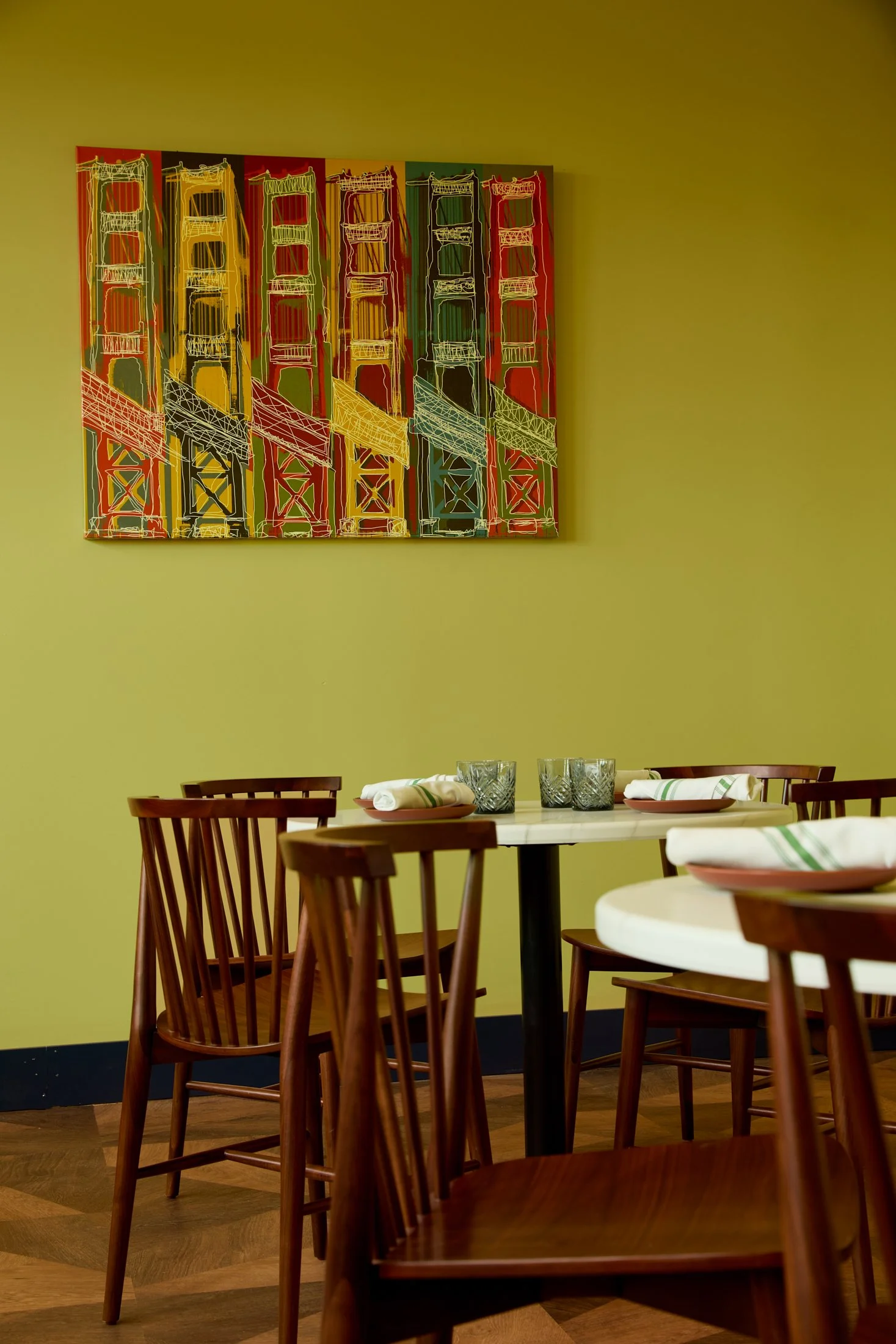 Interior of a restaurant with wooden chairs around white tables, set with napkins, glasses, and plates. A colorful art piece featuring a stylized bridge hangs on the yellow wall.