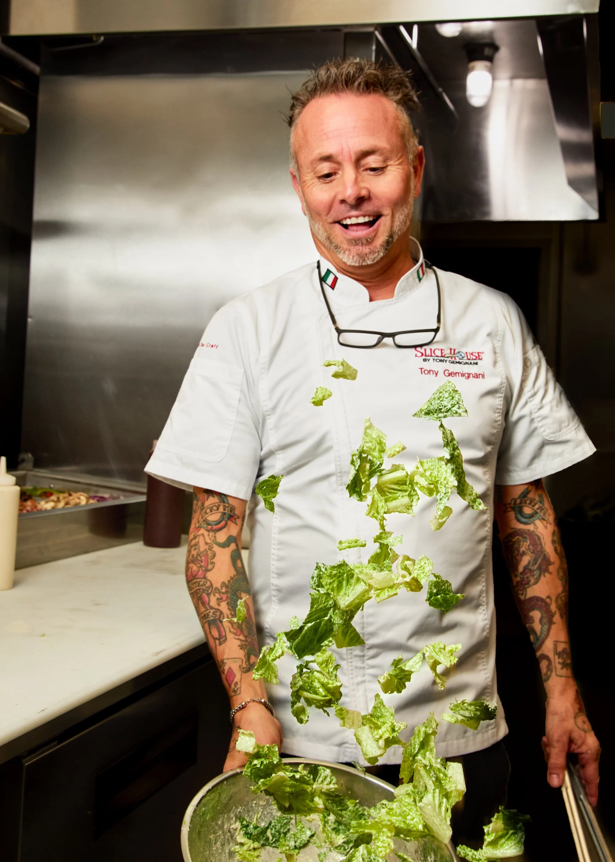 A man in a chef's coat tossing chopped lettuce in the air inside a commercial kitchen.