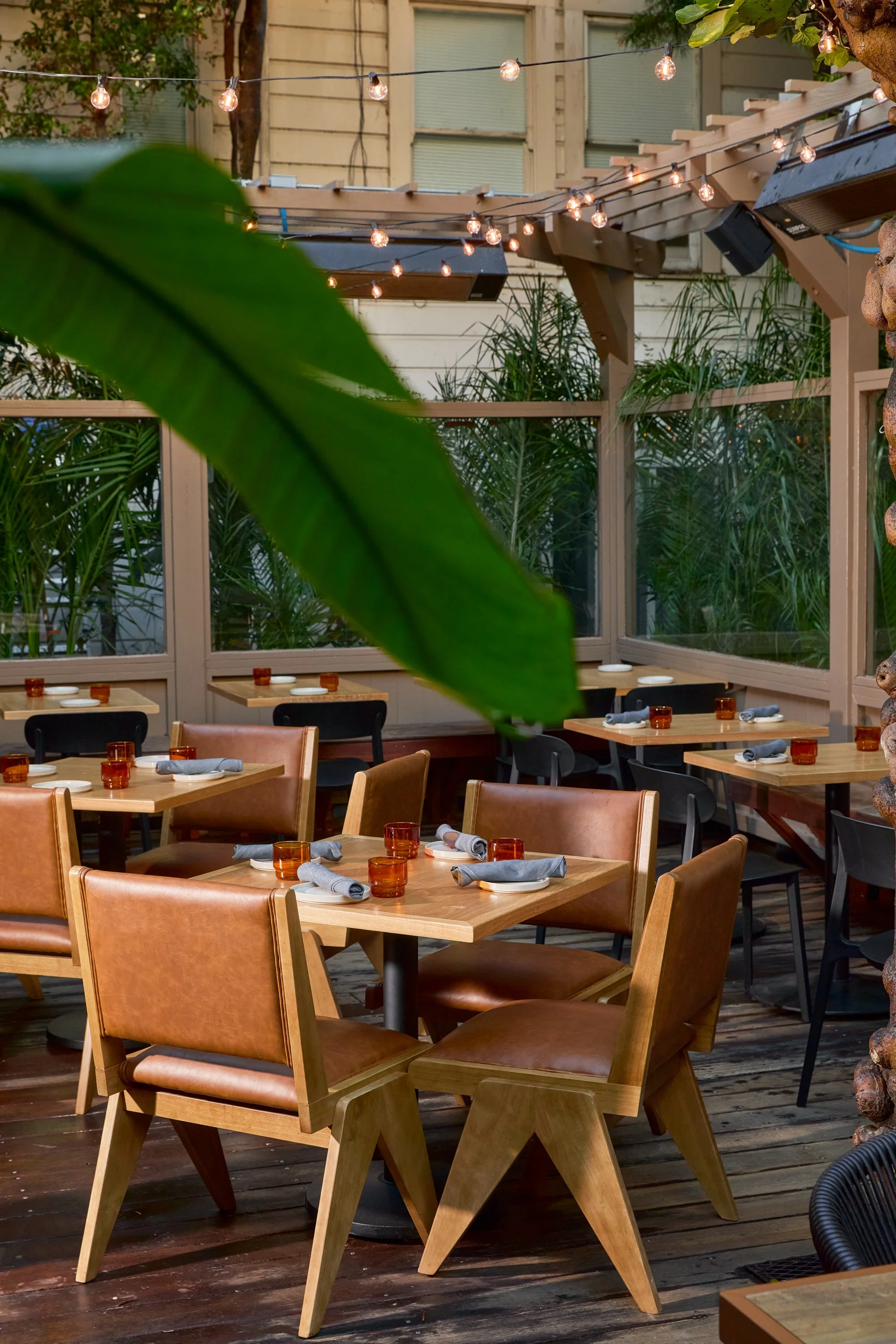 Empty outdoor restaurant patio with wooden tables, leather and plastic chairs, set with plates, napkins, and small orange glasses, and string lights overhead.