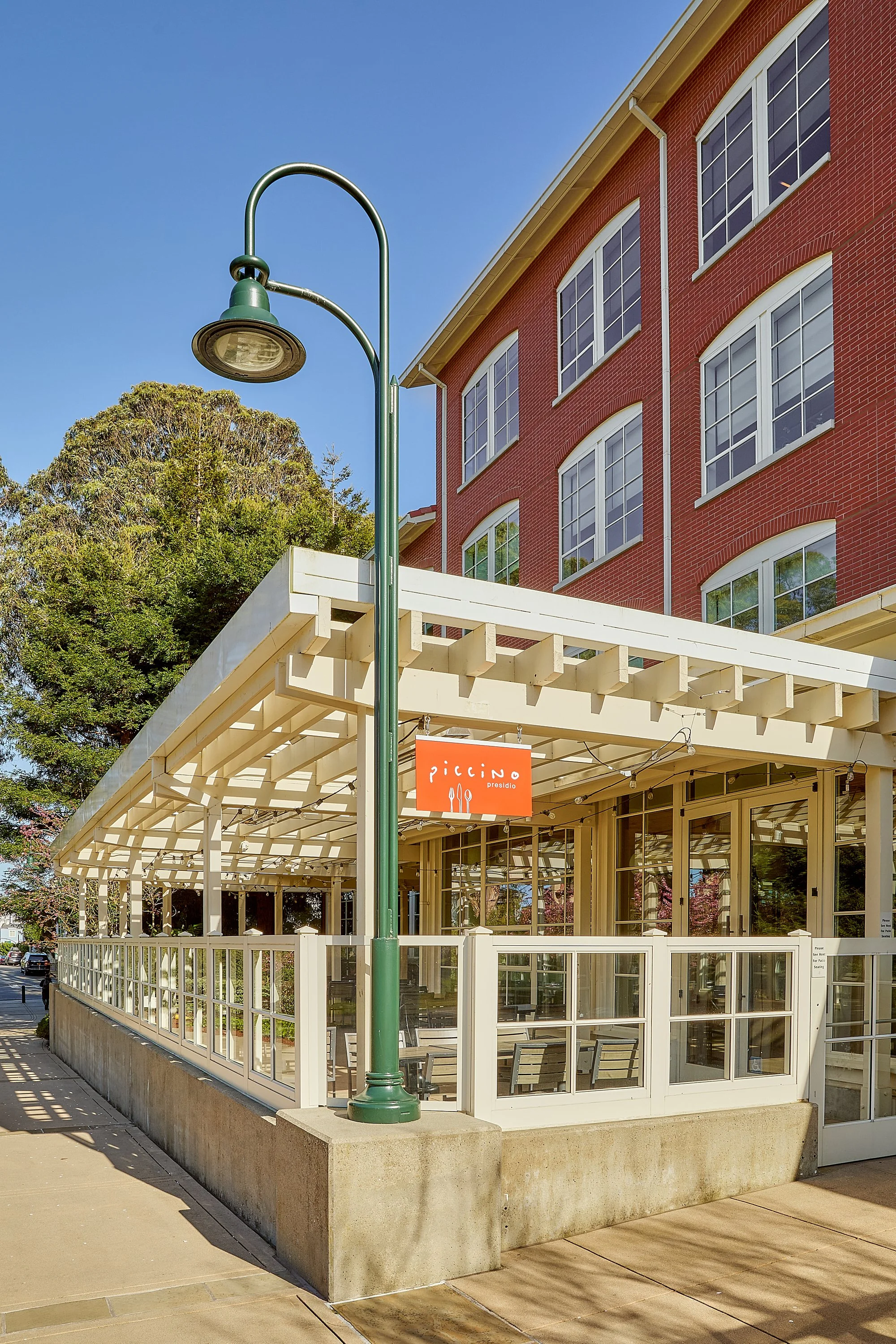 Street scene with a red brick building, a white fence, a green street lamp, and an orange and white restaurant sign reading 'Pizzino Presidio' under a clear blue sky.