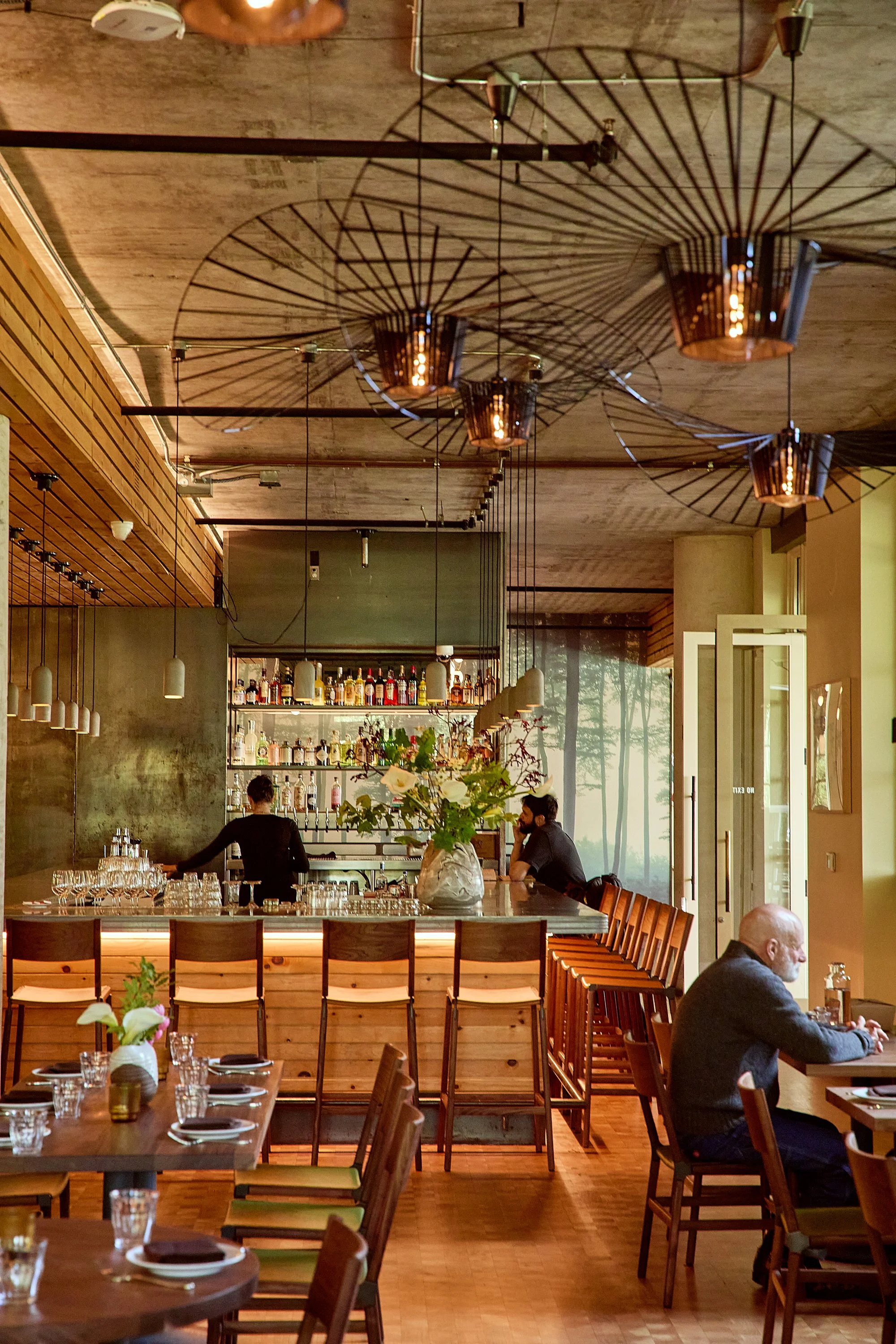 Interior view of a restaurant or bar with wooden decor, pendant and decorative lighting, a bar area with bottles, and several seated patrons at tables.
