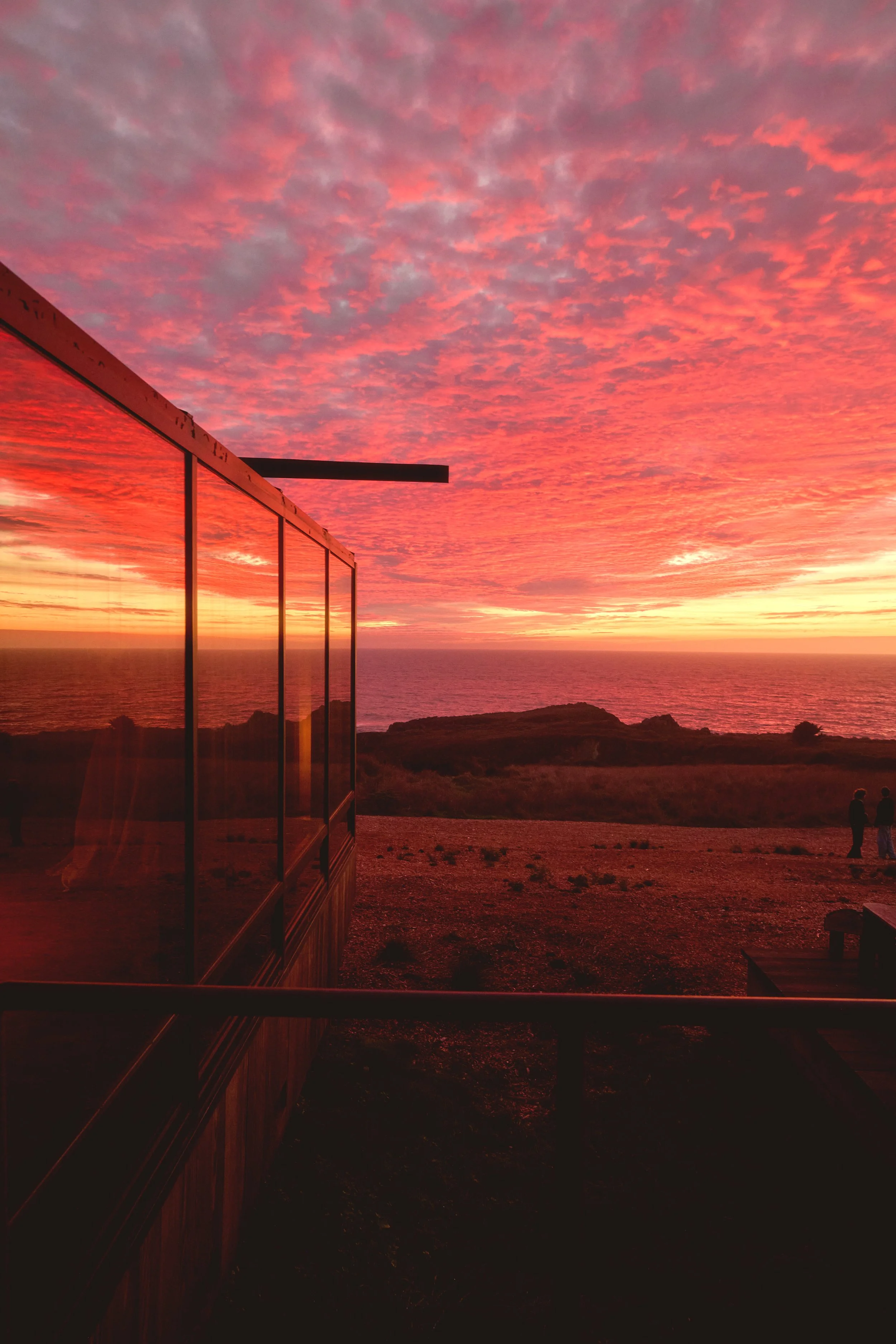 Sunset over the ocean with a colorful sky filled with pink, orange, and purple clouds. A glass building with reflections is on the left, and two people stand on the right on a grassy hill.