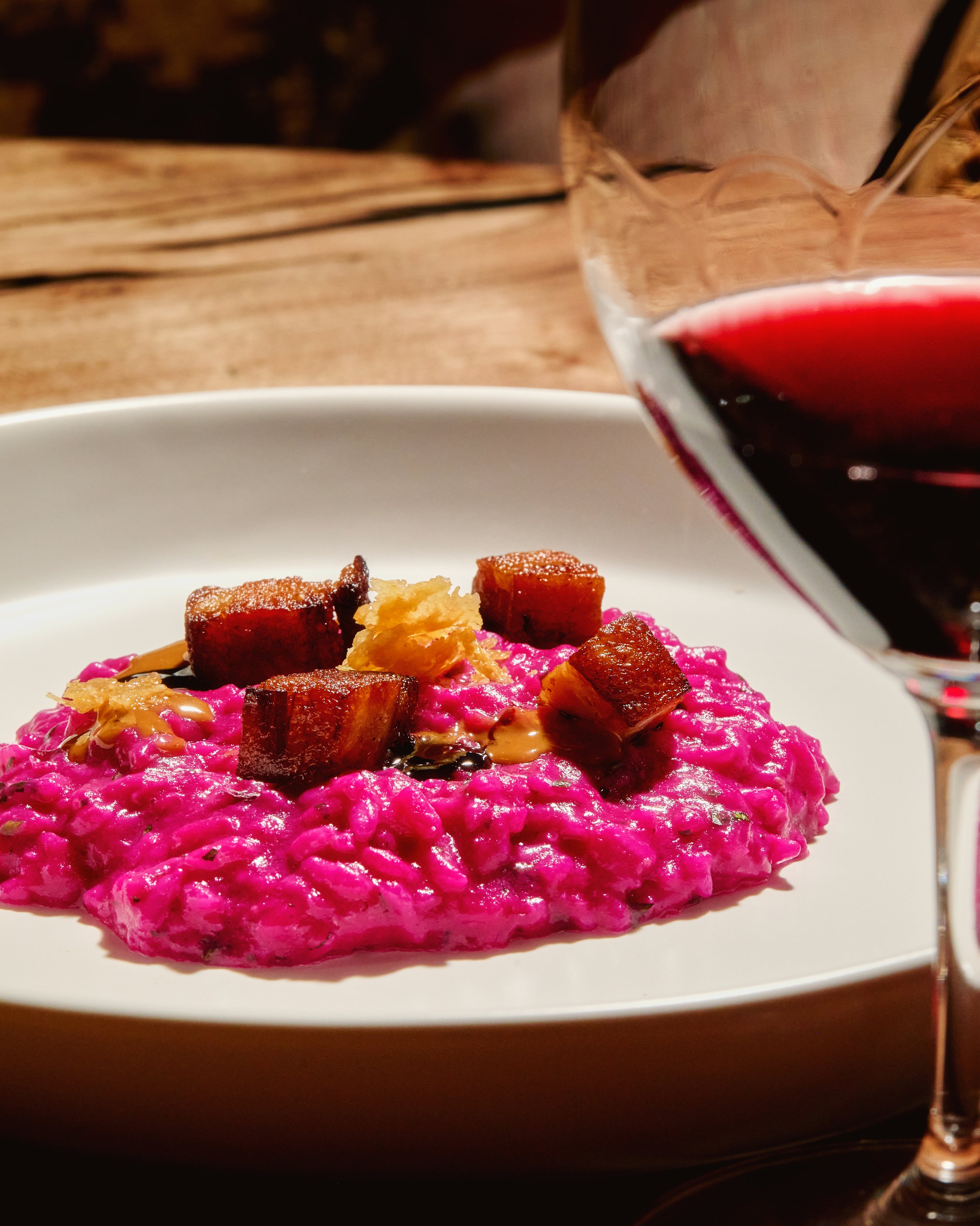 Pink beetroot risotto topped with pork belly pieces and a drizzle of sauce, served on a white plate with a glass of red wine in the foreground.