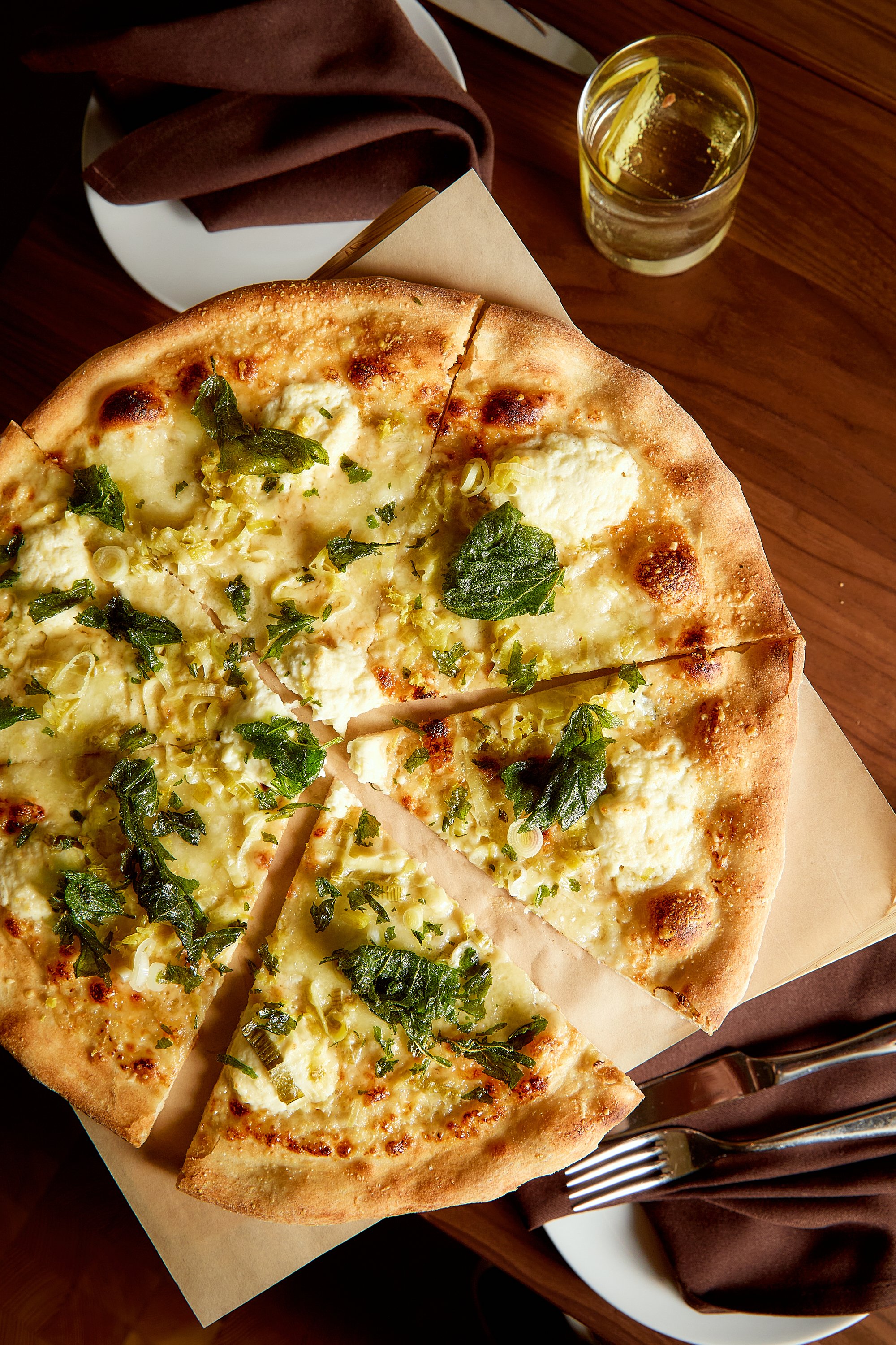 A pizza with cheese, herbs, and green onions on a brown crust, partially sliced on a tray. A glass of water and a napkin with utensils are on a wooden table.